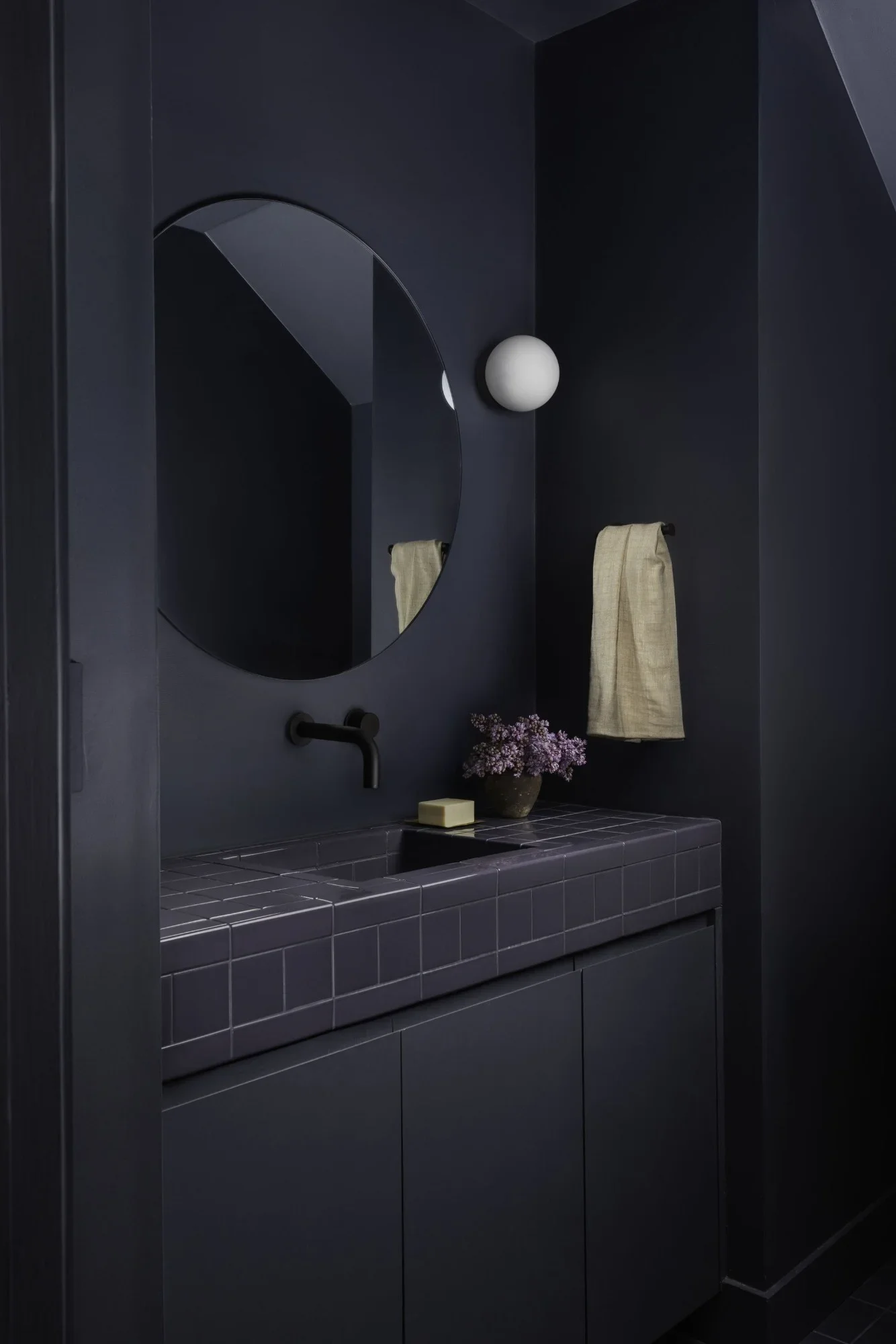 Moody powder room with dark navy walls, a round frameless mirror, matte bronze wall-mounted faucet, and a tiled vanity counter and integrated sink in deep navy.