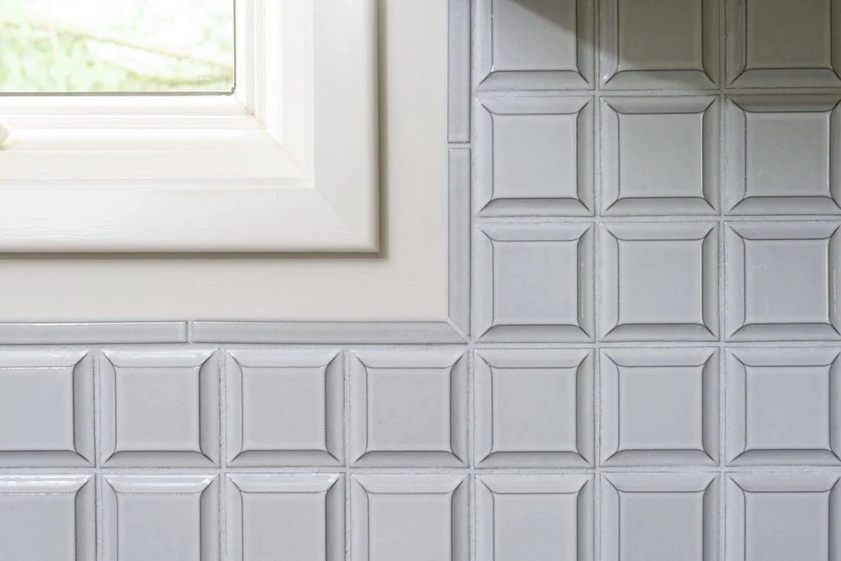 Close-up of pale blue cushion-edge ceramic wall tile with dimensional relief pattern next to white window trim