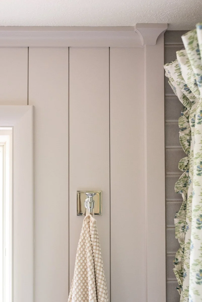 Detail of painted vertical shiplap wall paneling with crown molding, polished chrome robe hook, and ruffled floral shower curtain