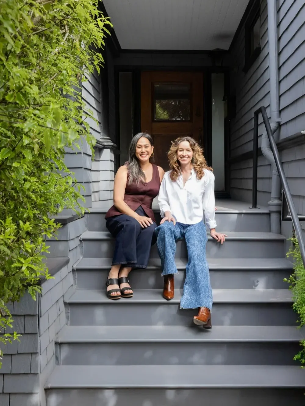 Two women sitting on the front steps of a house, smiling at the camera. One woman has long black hair, wearing a sleeveless burgundy top and dark pants. The other has curly red hair, wearing a white blouse and blue bell-bottom jeans. The house has gray siding and black railing, with greenery on the sides.