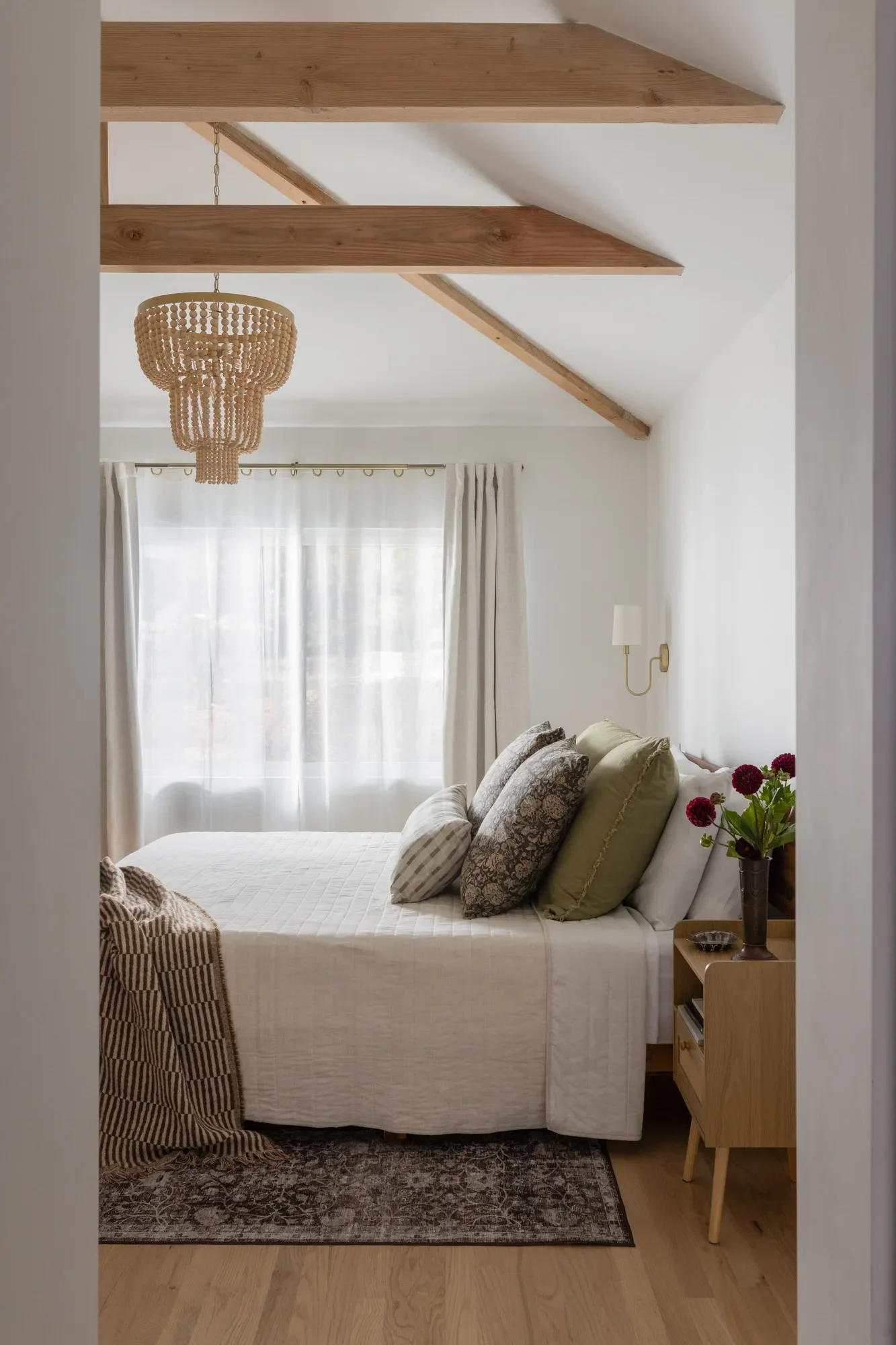 Bedroom with vaulted ceiling, exposed wood beams, layered linen bedding, and wood-bead chandelier in Carmichael home designed by Studio VAE, San Francisco interior designer.