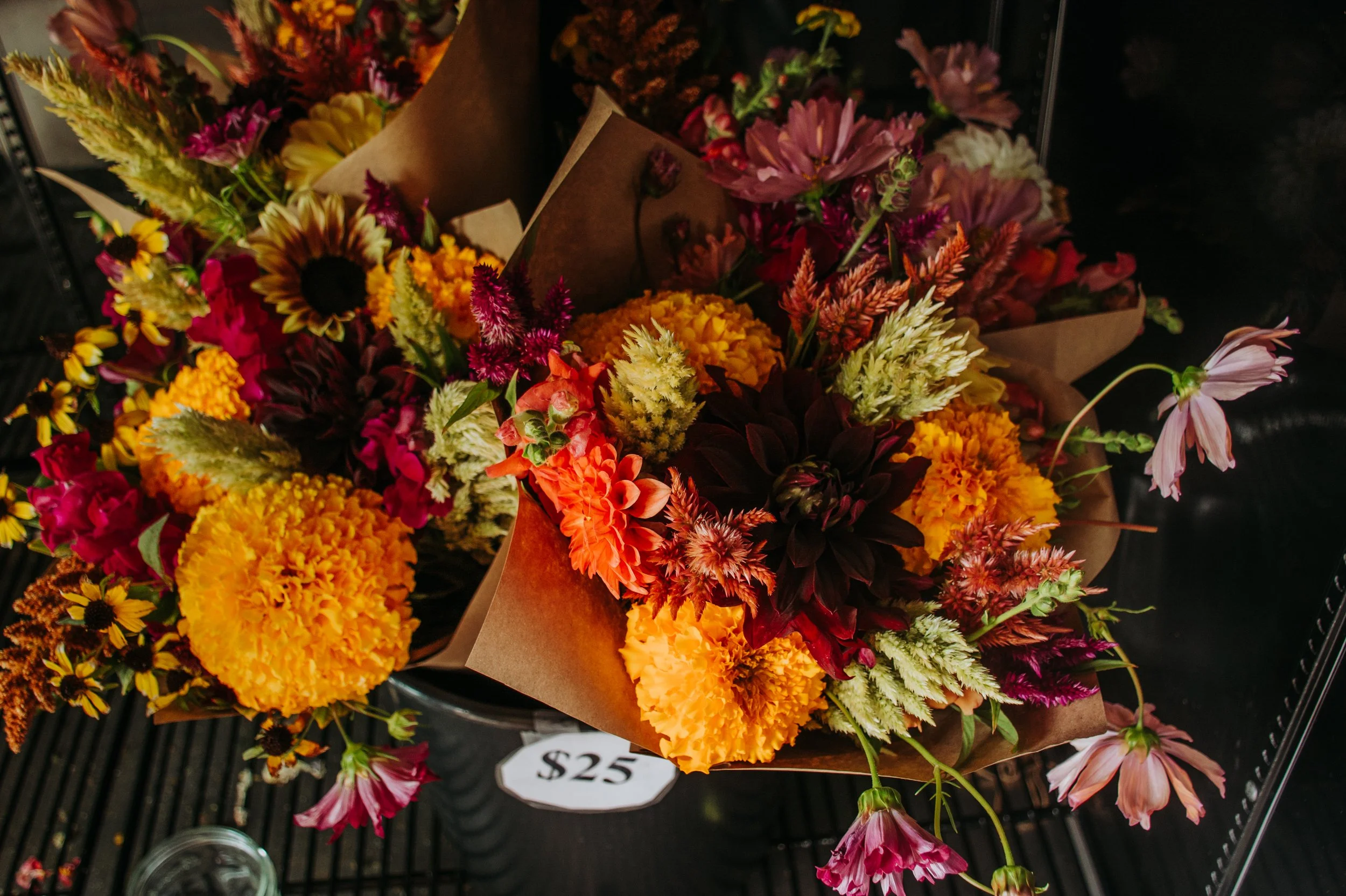 Colorful bouquet of various flowers including marigolds, sunflowers, zinnias, and roses, wrapped in brown paper, with a price tag of $25 placed underneath on a black wire shelf.