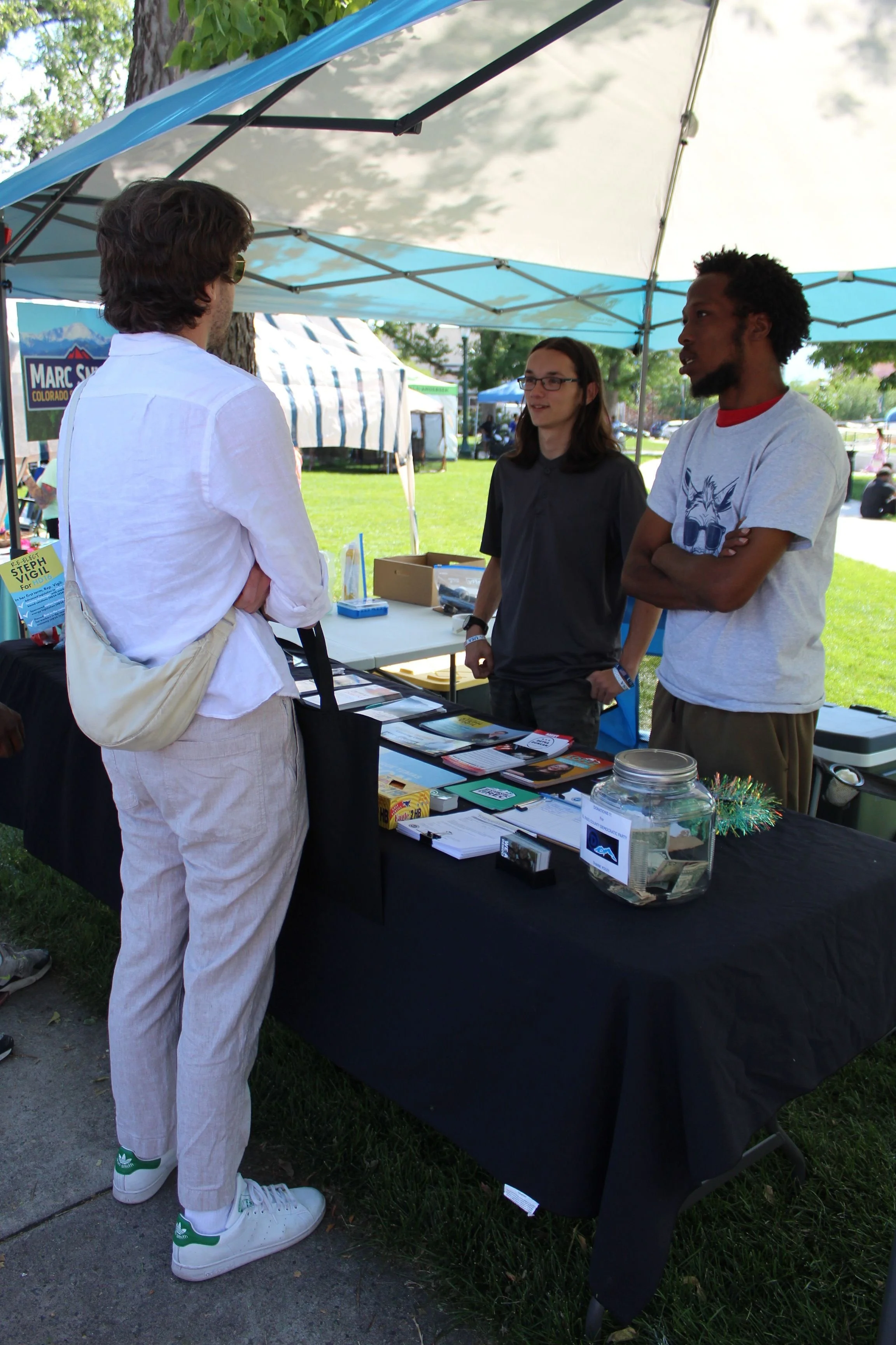 Outdoor market stall with three people interacting under a canopy. The table displays books, pamphlets, and a donation jar.