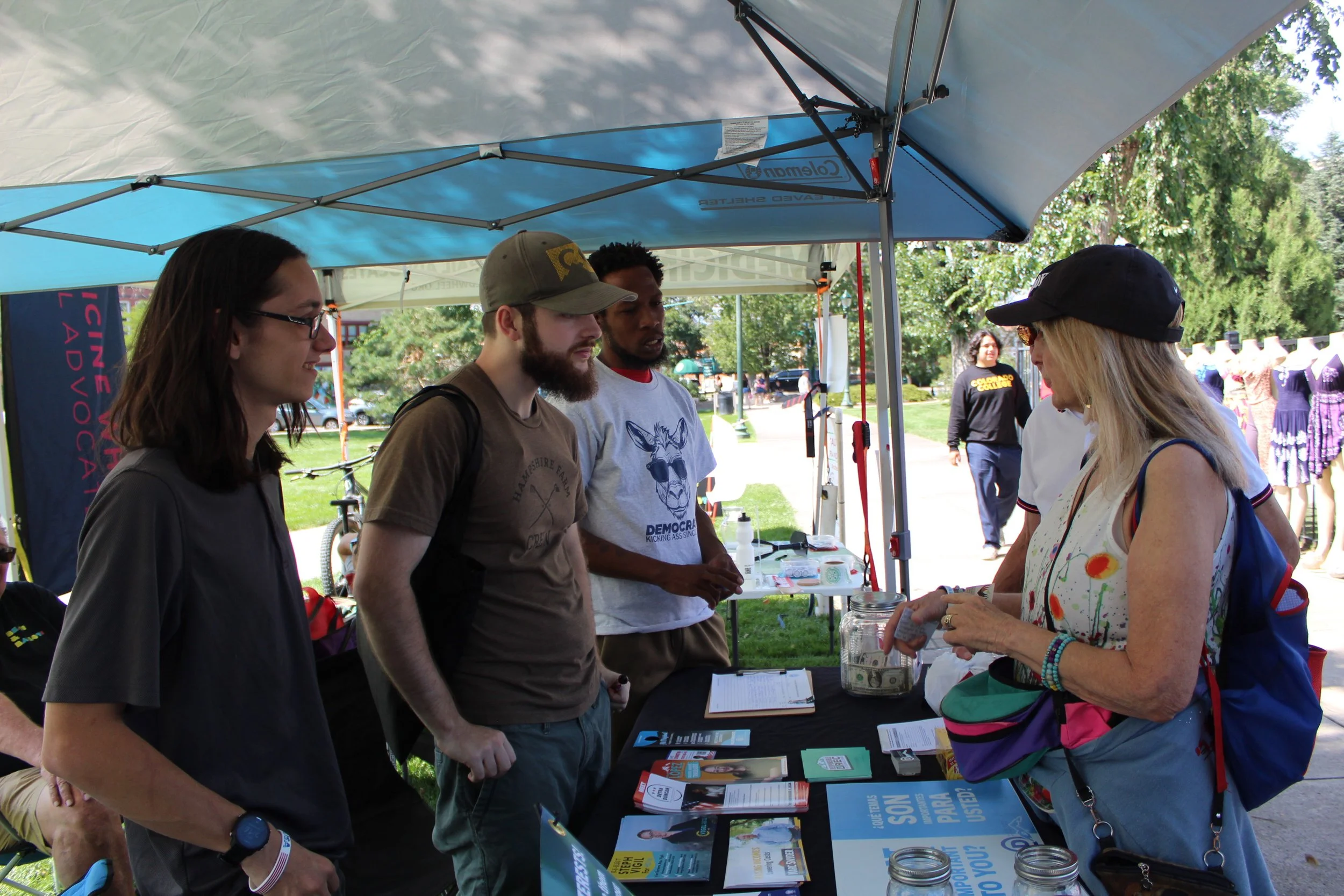 People at an outdoor advocacy booth under a tent, discussing materials on a table.