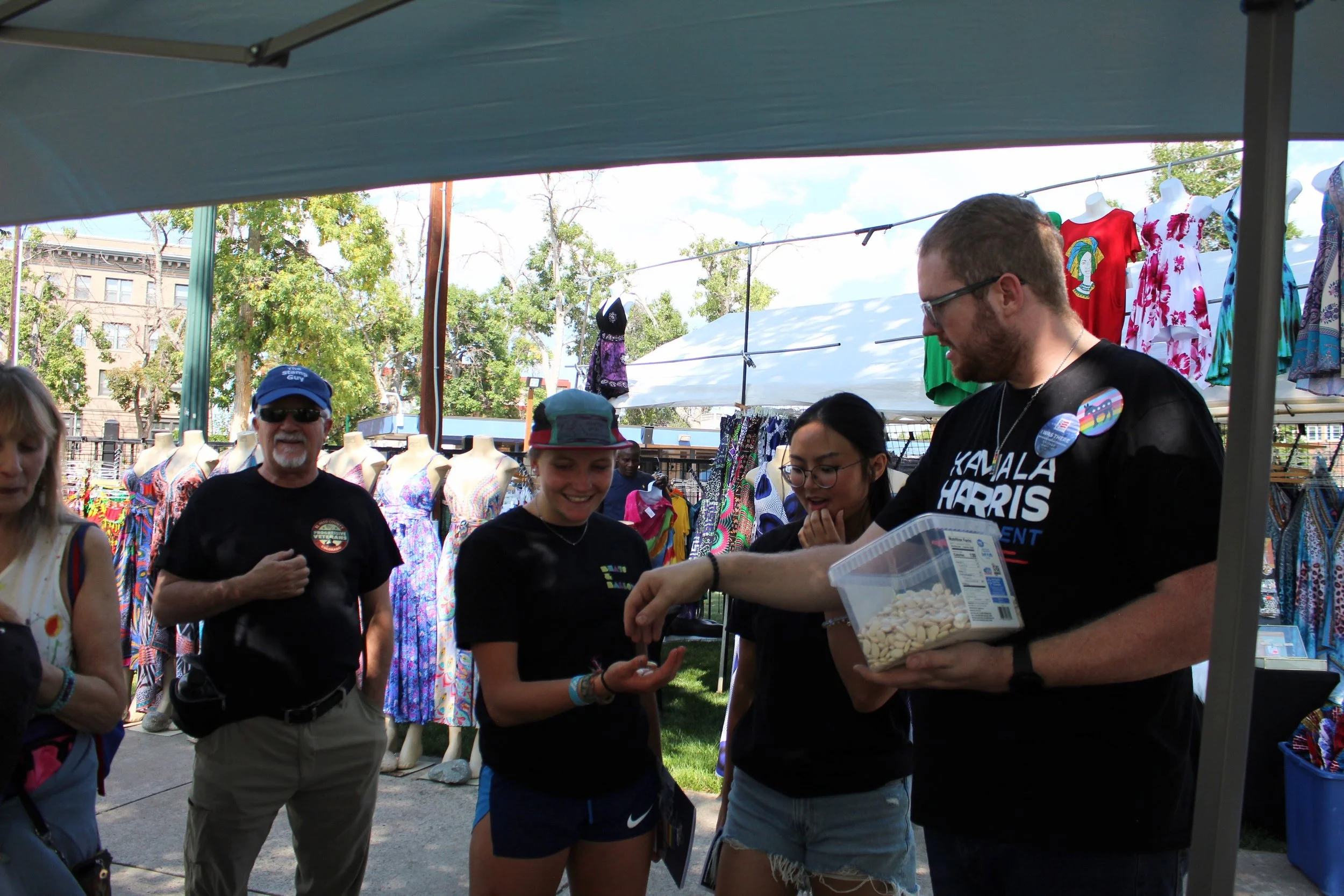 Group of people in outdoor market with colorful dresses and a man holding a box of beans.
