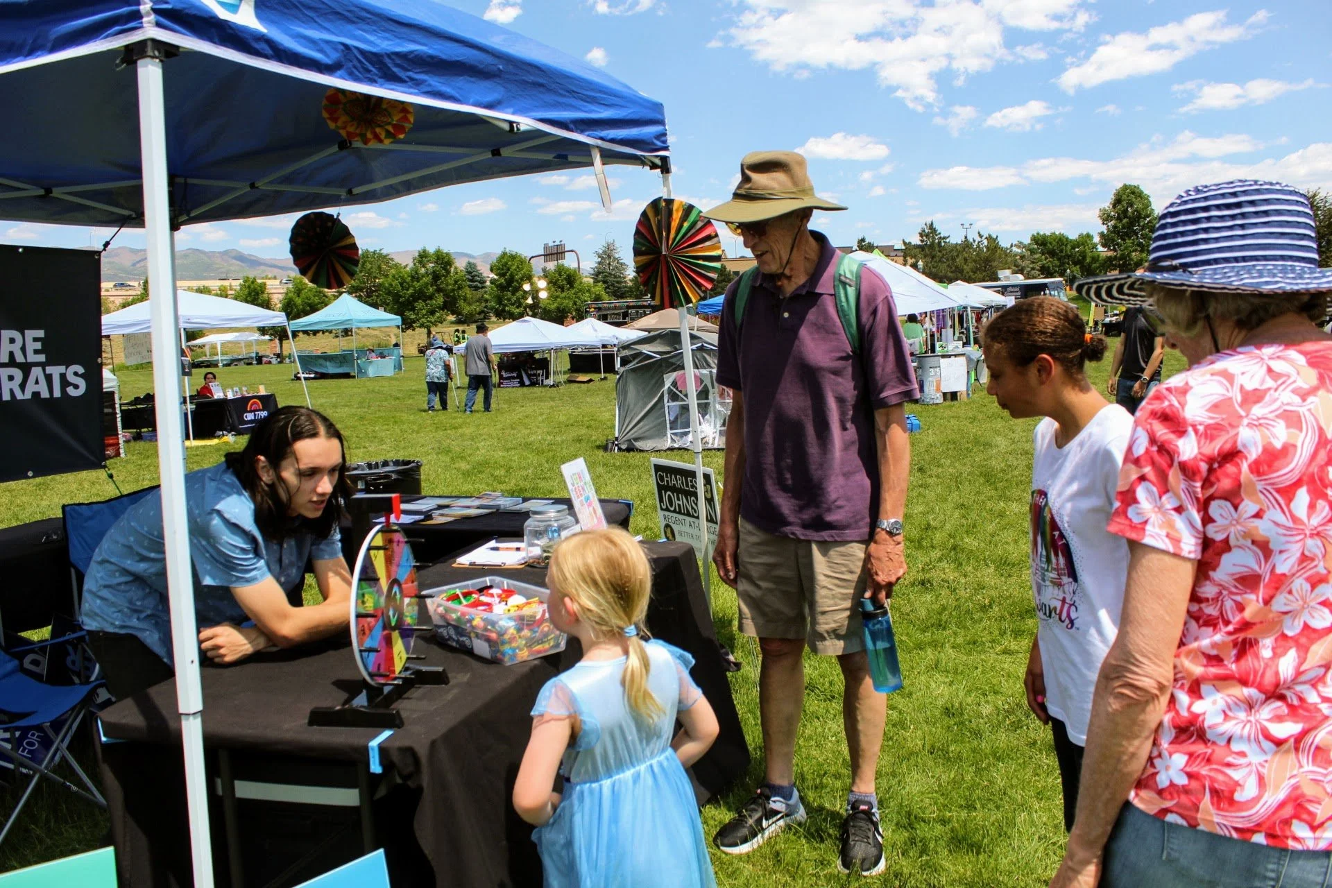 Outdoor fair with people gathered at a booth under a blue tent, colorful spinning wheel, and candy bowl on a table.