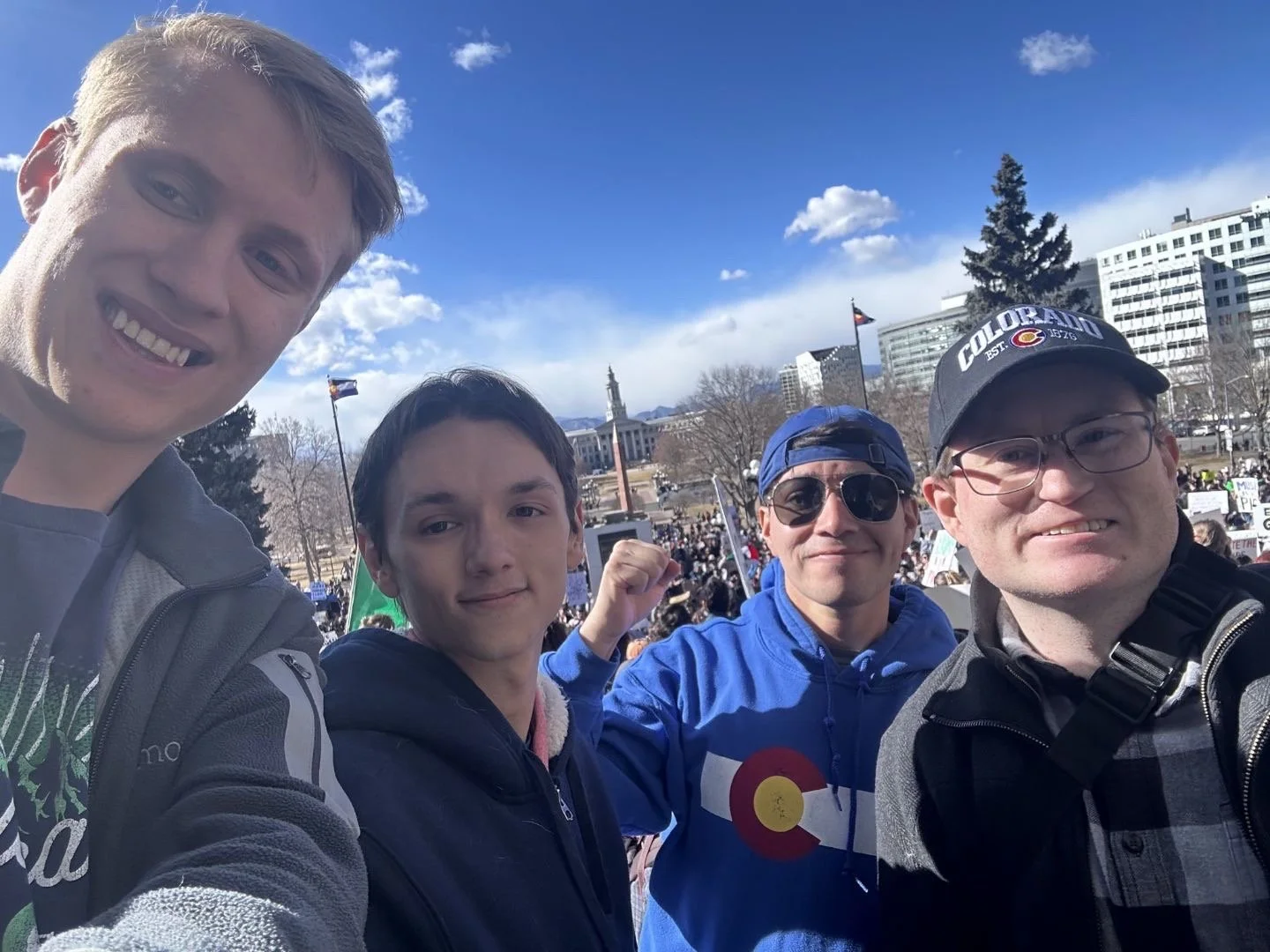 Group of four people taking a selfie outdoors during a public event or protest, with a crowd and buildings in the background and Colorado flags visible.