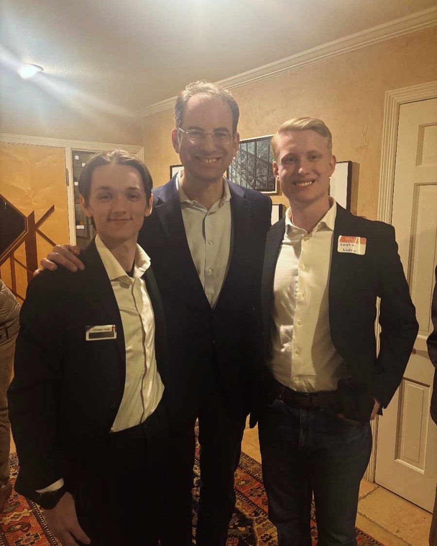 Three men in suits posing for a photo indoors, wearing name tags; one in the center with glasses and the other two smiling on either side.