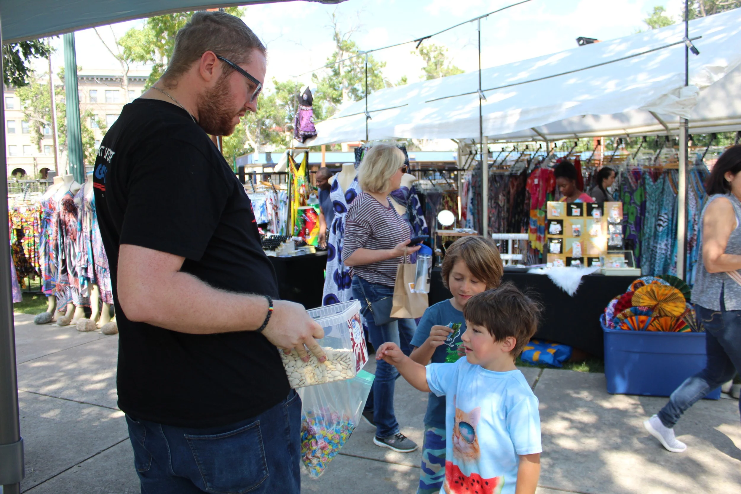 Vendor interacting with children at outdoor market, offering items from a plastic container, with clothing stalls and people in the background.