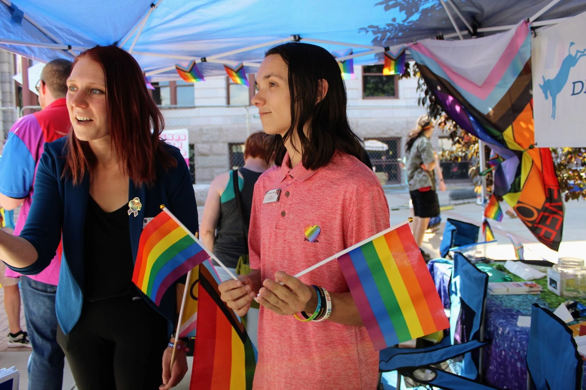 People under a tent holding rainbow pride flags at an LGBTQ+ event.