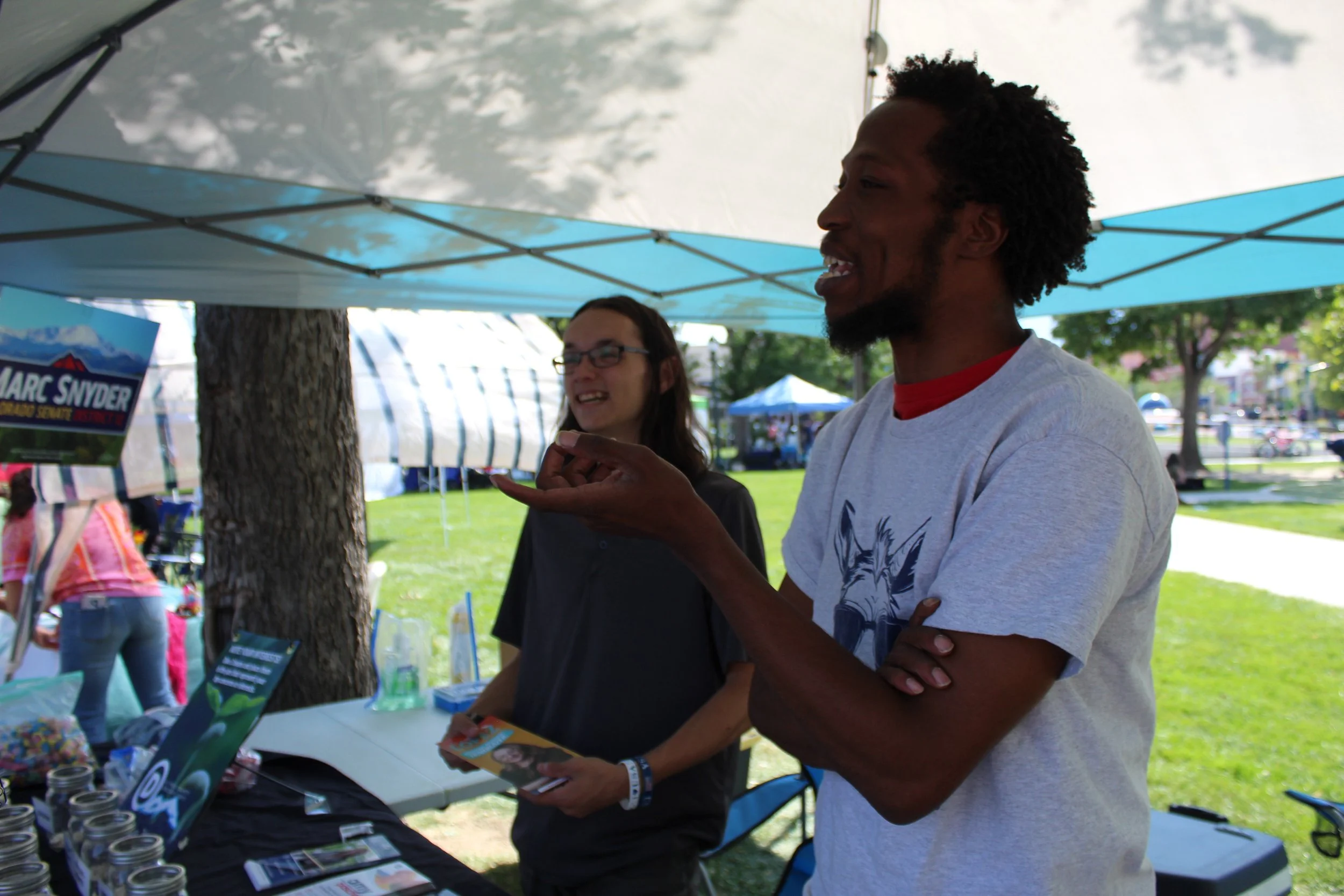 Two people at a market booth under a tent, with promotional materials and jars on display, trees and a park visible in the background.