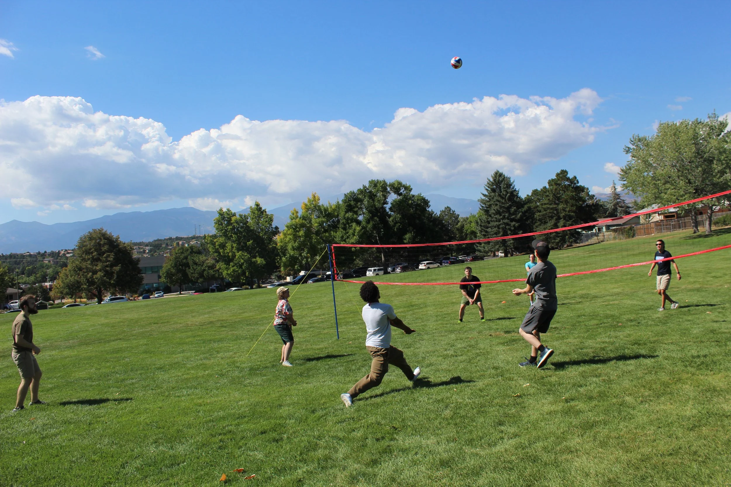People playing volleyball on grassy field with trees and mountains in background.