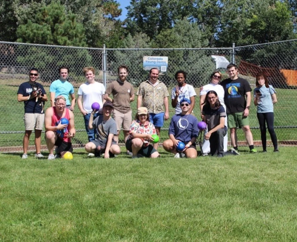 Group of people posing with balls in a park setting with a chain-link fence and sign in the background.