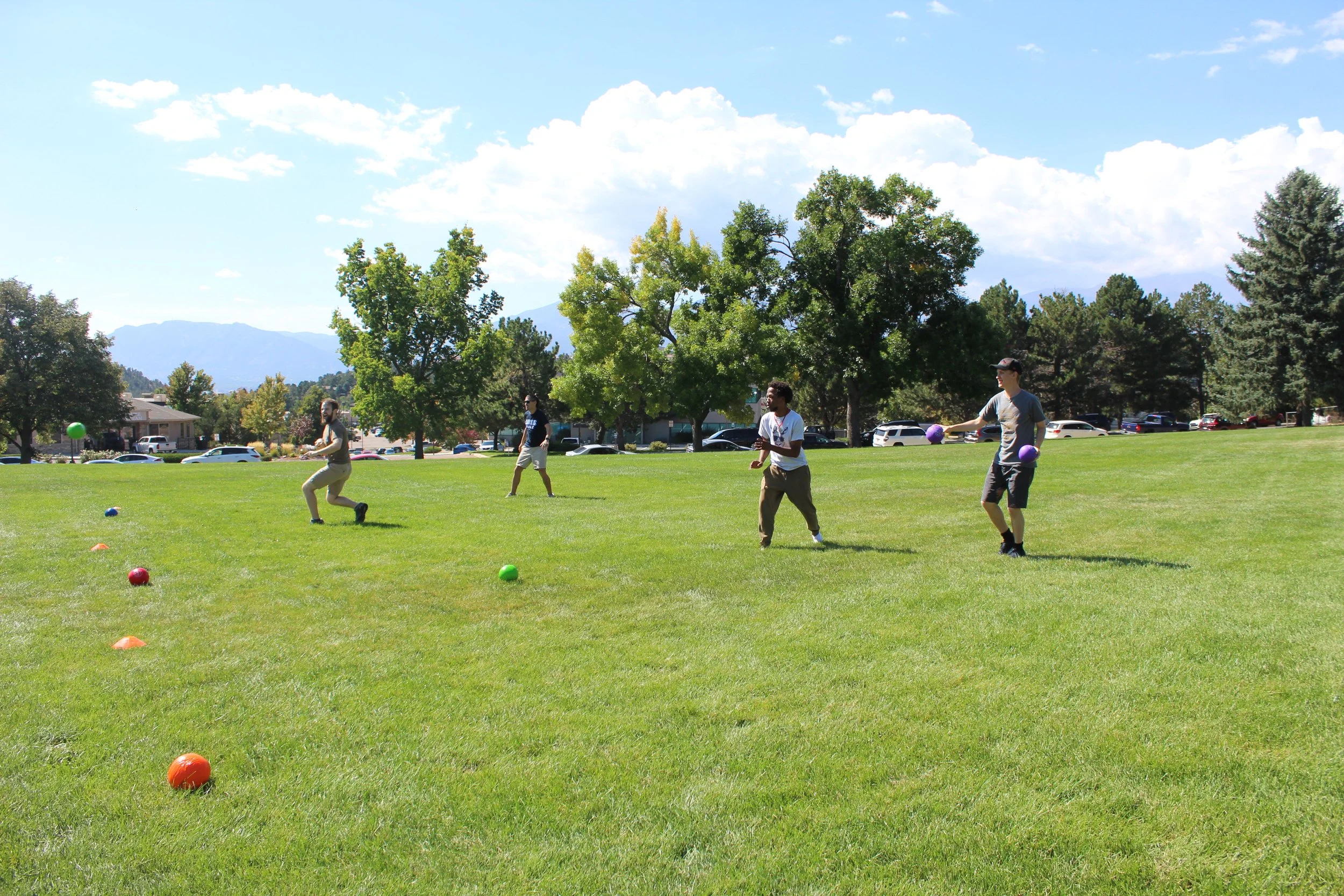 People playing dodgeball on a grassy field with trees and mountains in the background.