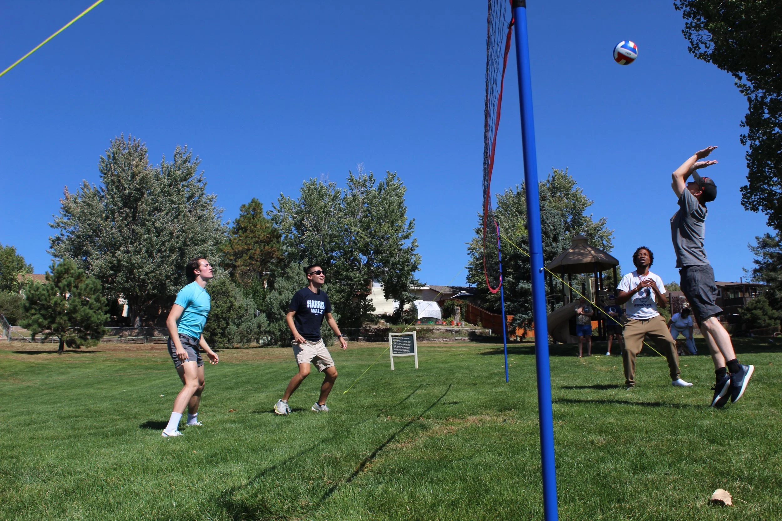 Group of people playing volleyball outdoors on grass field with blue sky and trees in the background.