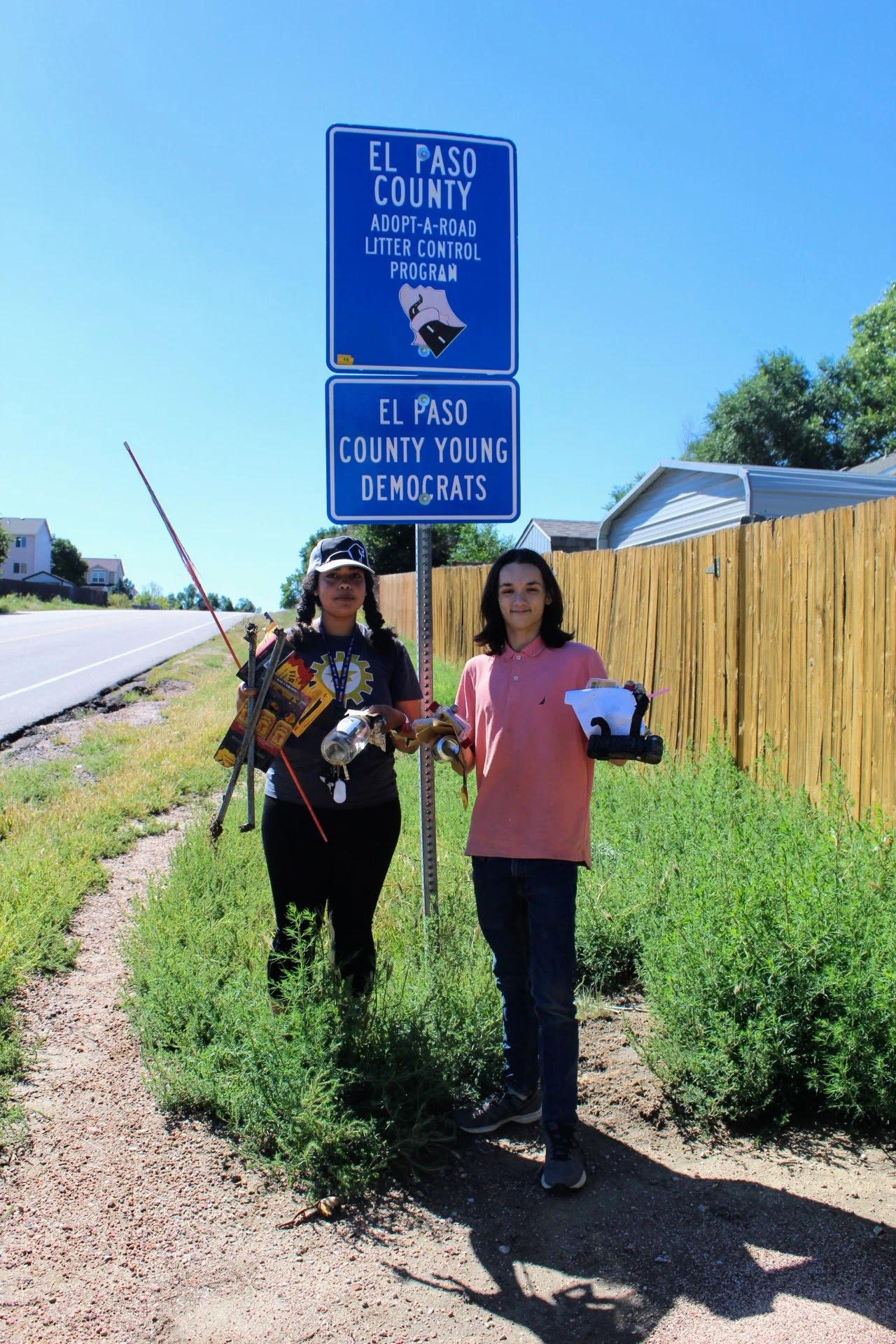 Two people standing near an El Paso County sign for the Adopt-a-Road Litter Control Program and El Paso County Young Democrats.