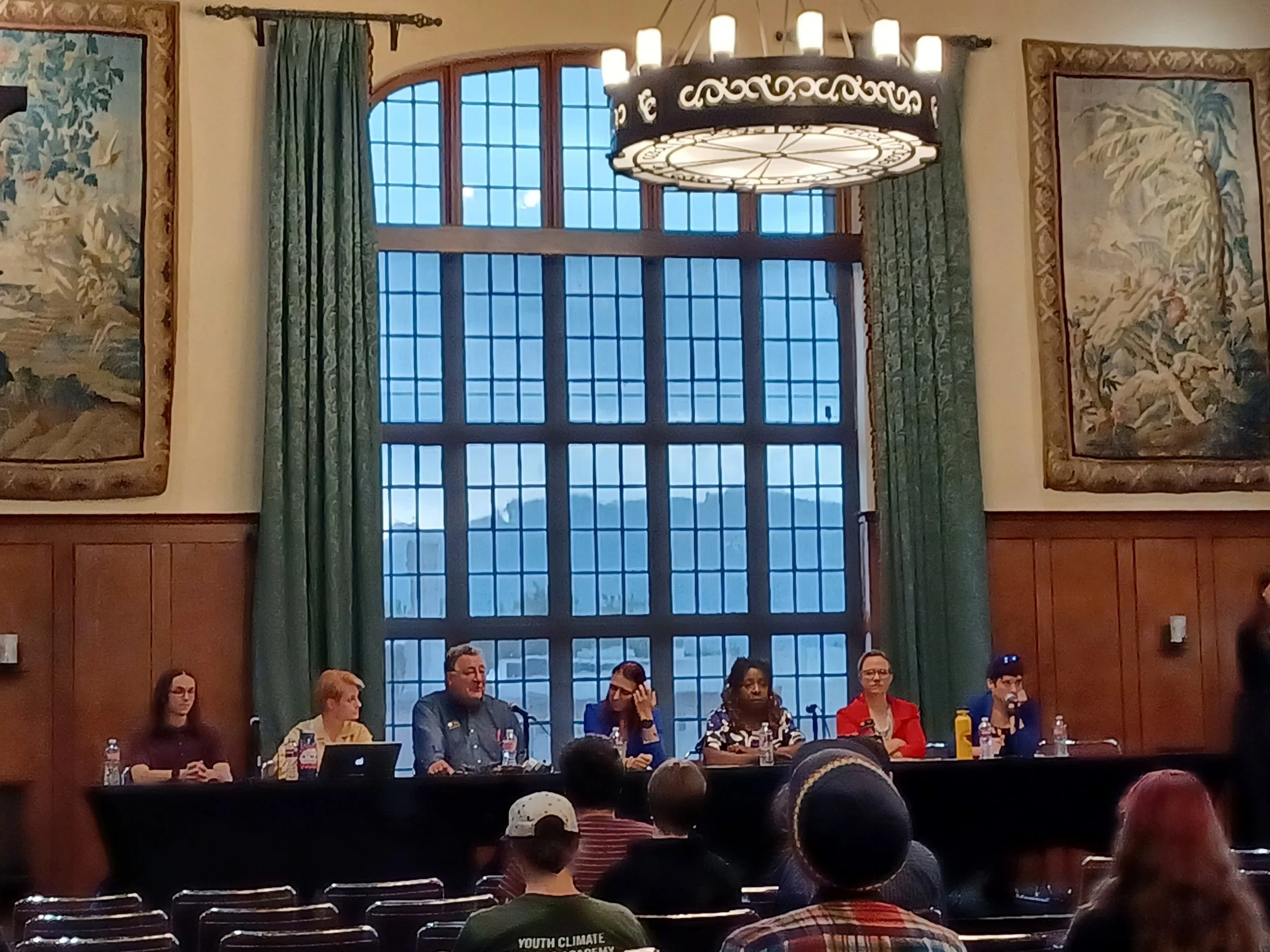 Panel discussion in a historic room with six speakers at a long table, vintage decor, and audience in foreground.