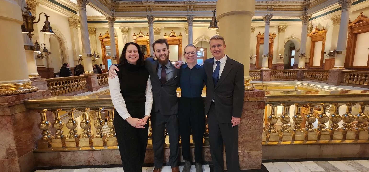 Group of four people posing in a grand interior with columns and decorative railings.