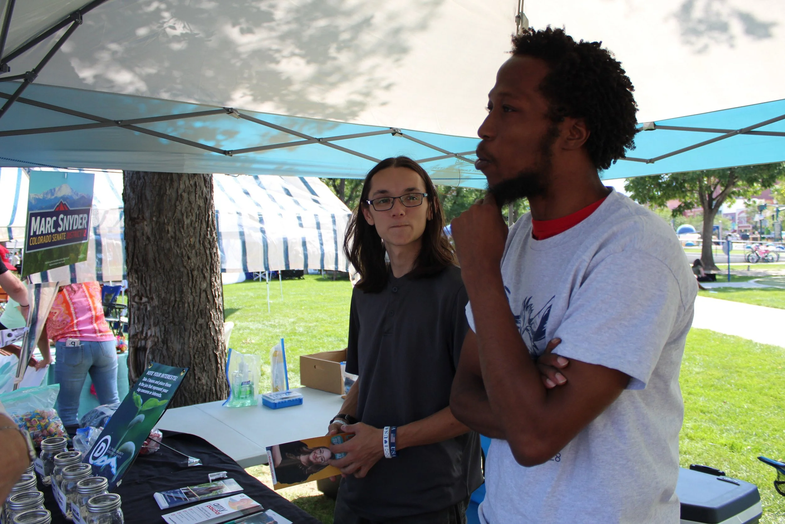 Two people talking under a tent at an outdoor event with promotional materials and a political campaign sign in the background.