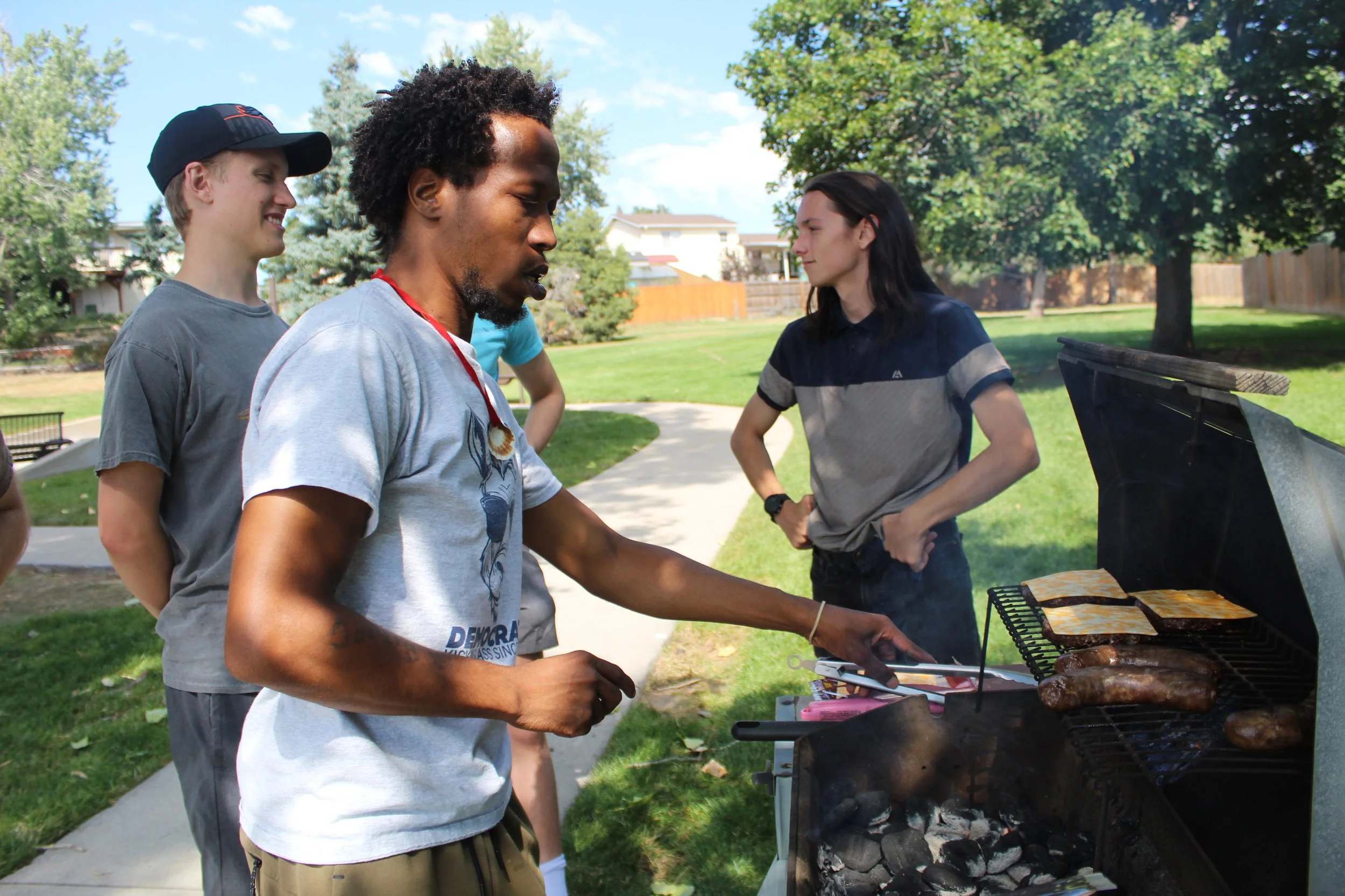 Three people barbecuing in a park, one grilling sausages and cheese on a charcoal grill.
