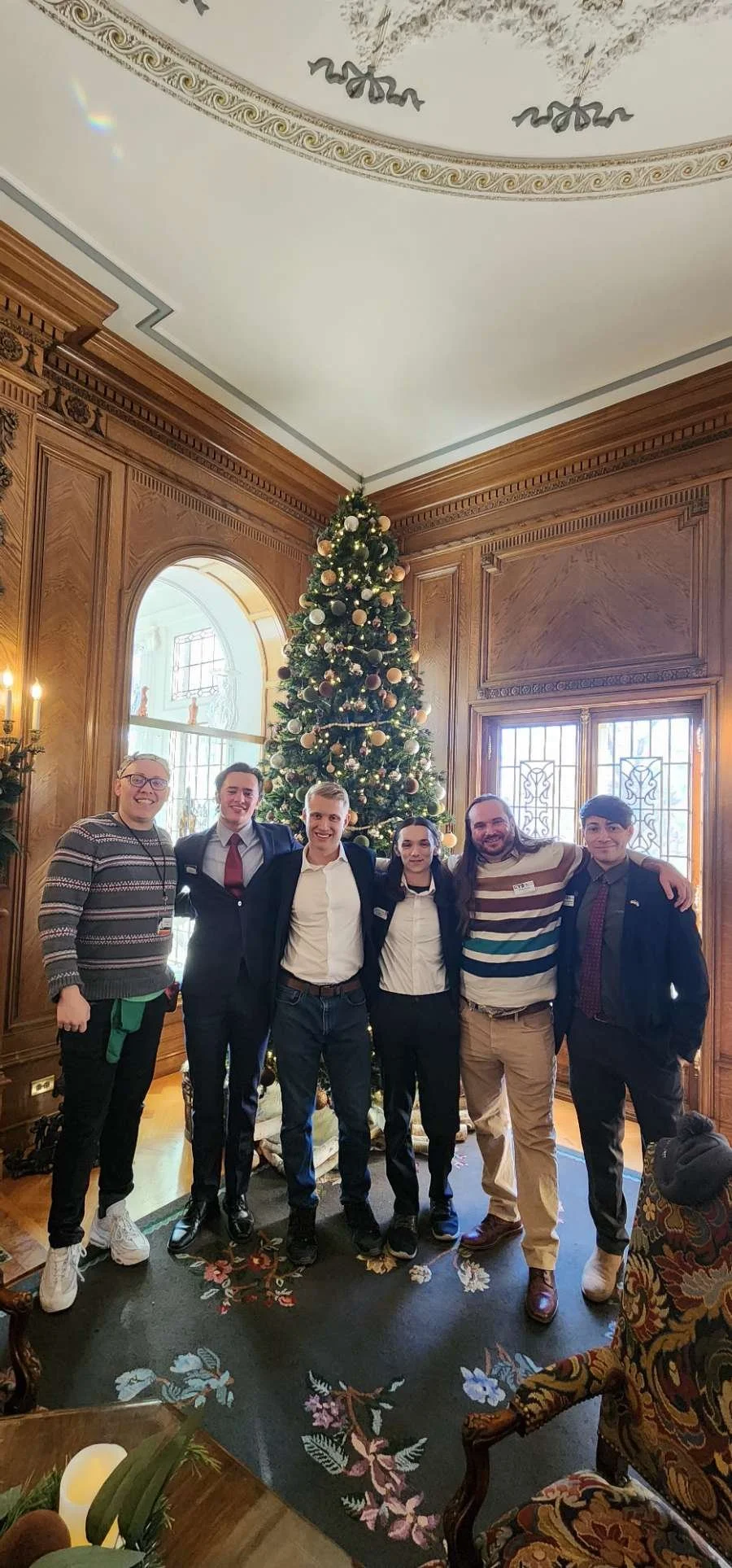 Group of six people posing in front of a decorated Christmas tree in an ornate room.