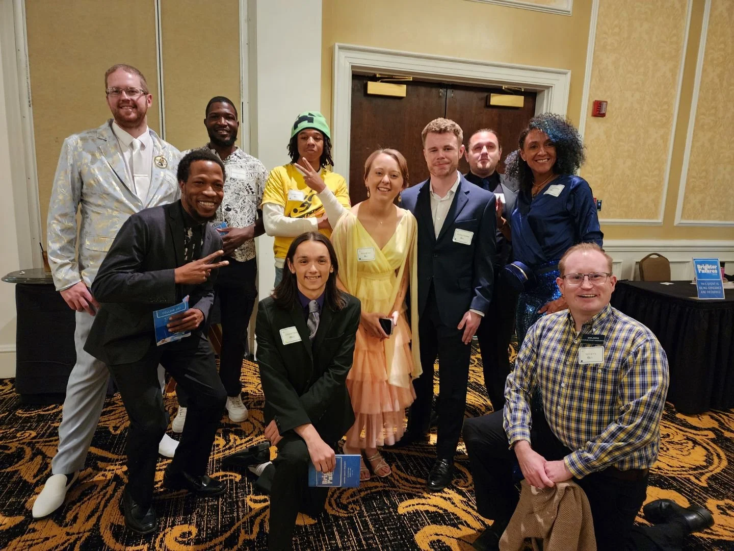 Group of nine people posing at a formal event with a carpeted floor and elegant decor. They are wearing a mix of formal and casual attire, some holding programs or brochures. The backdrop includes double doors and a table with a sign.