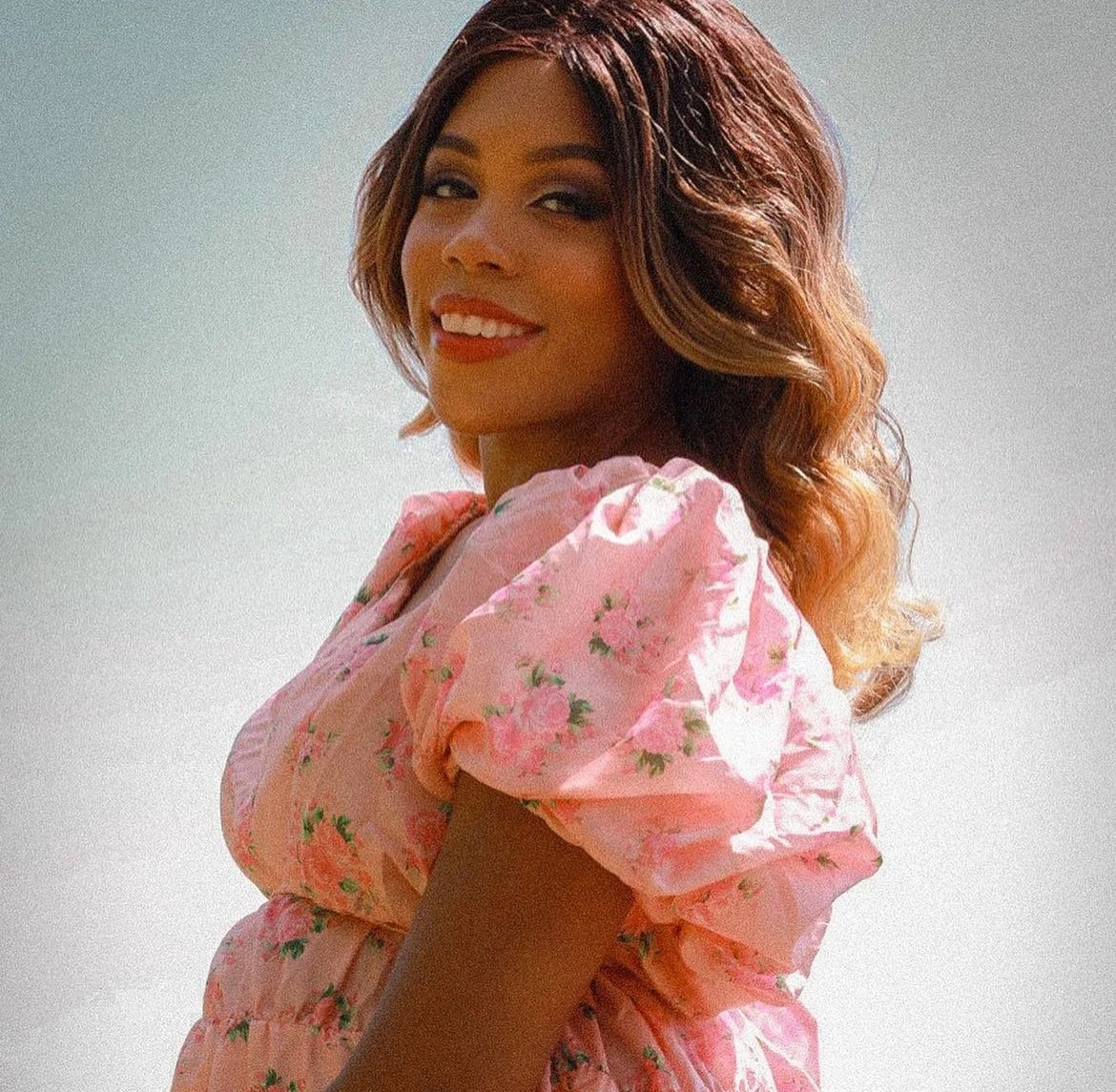A woman with wavy hair and red lipstick, smiling at the camera, wearing a pink floral dress against a plain background.