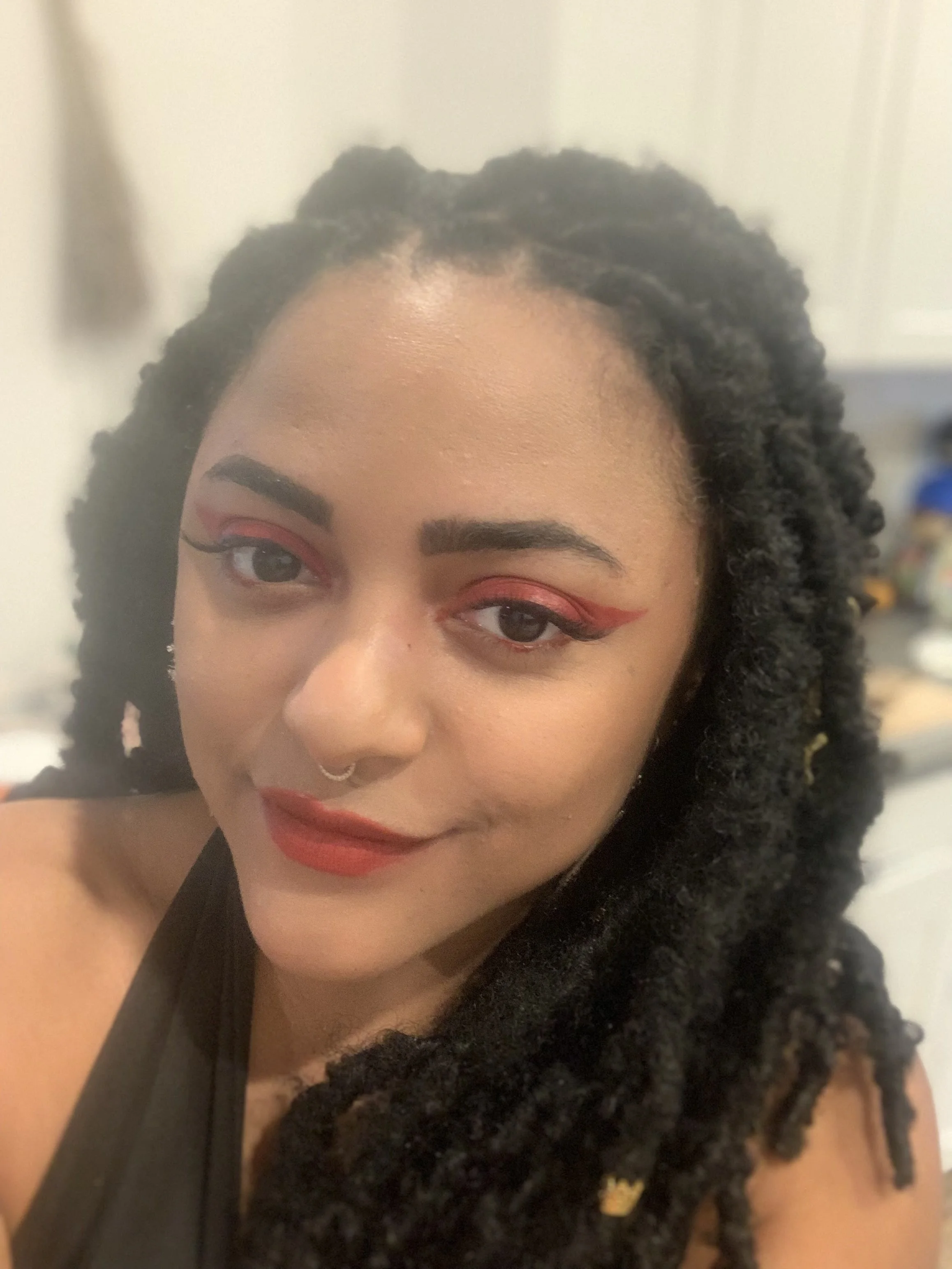 A woman with dark curly hair styled in twists, wearing red makeup and a septum piercing, smiling at the camera.