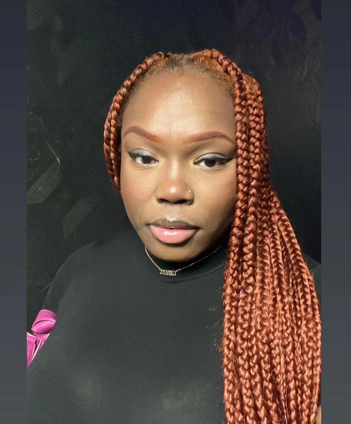 A woman with long, braided red hair looking at the camera with a neutral expression, wearing makeup, a nose piercing, a black top, and a gold necklace.