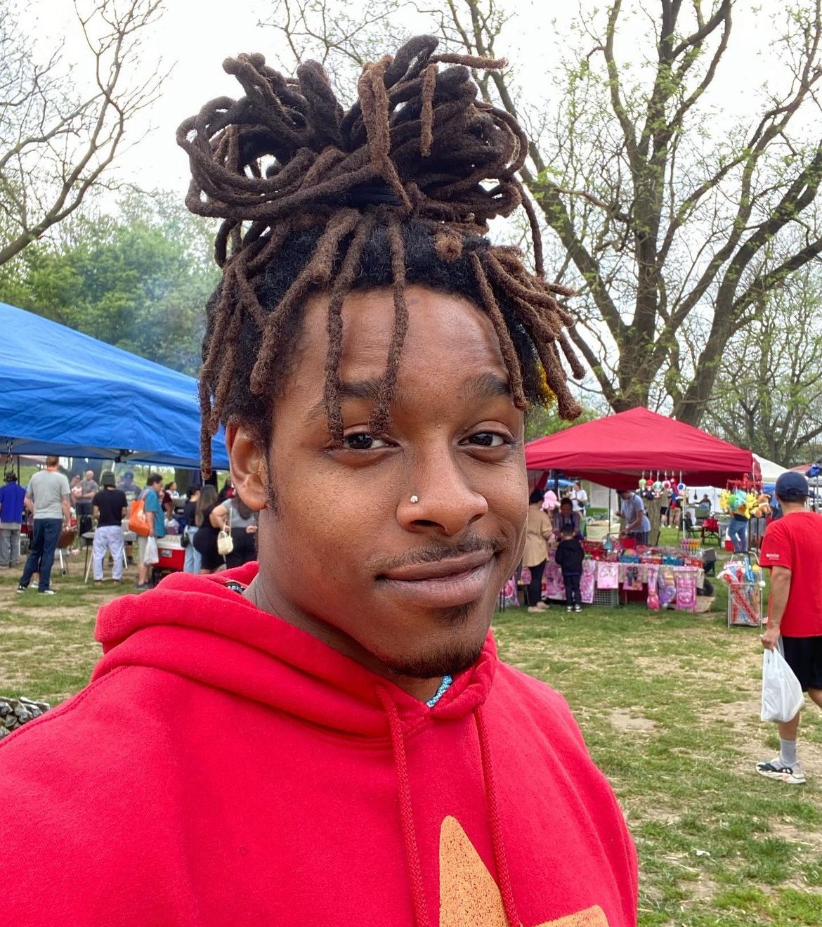 A young man with brown dreadlocks tied in a bun, wearing a red hoodie, in an outdoor setting with tents and people at a fair or market.