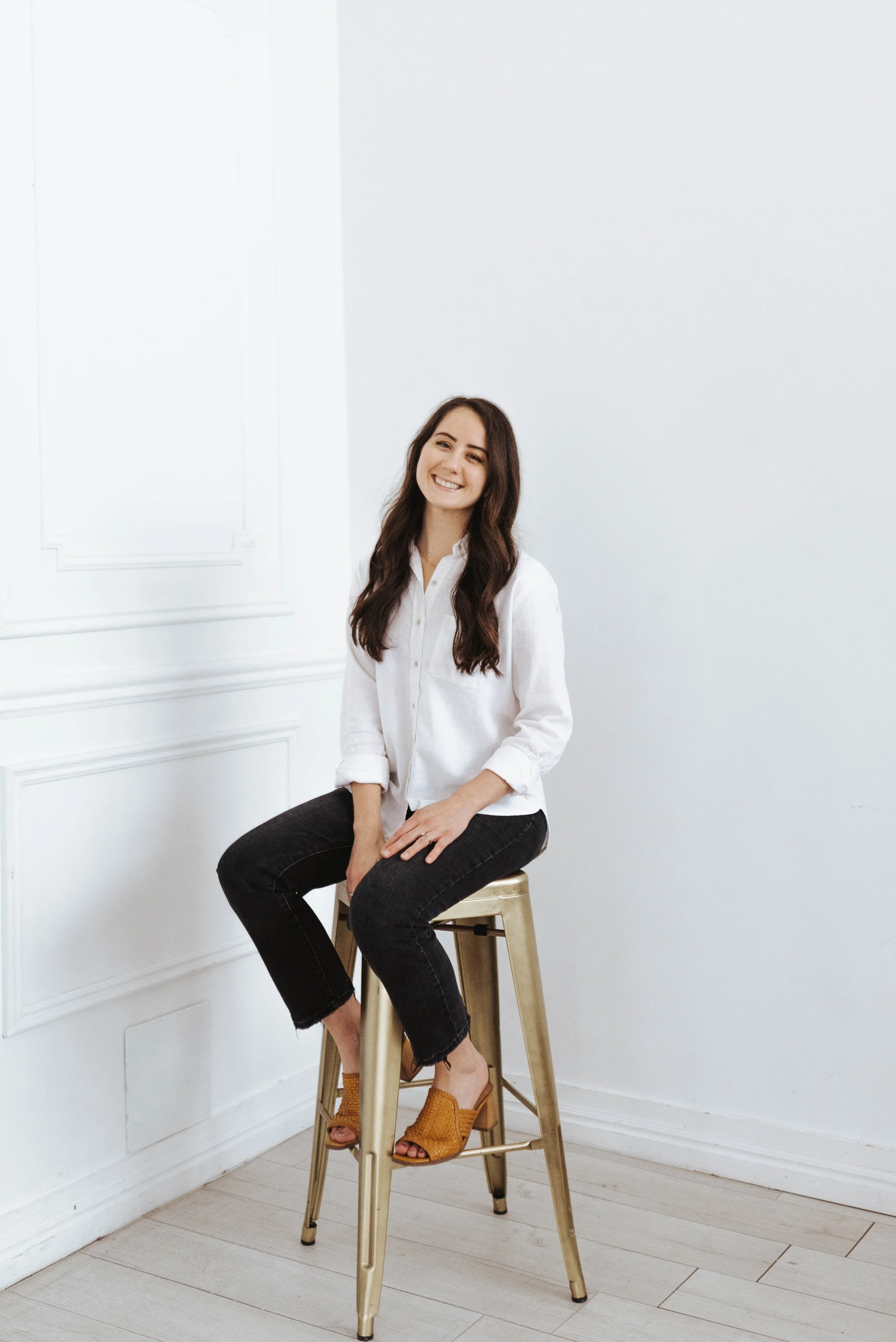 A young woman with long dark hair smiling while sitting on a wooden stool in a bright, white room with minimal decor.