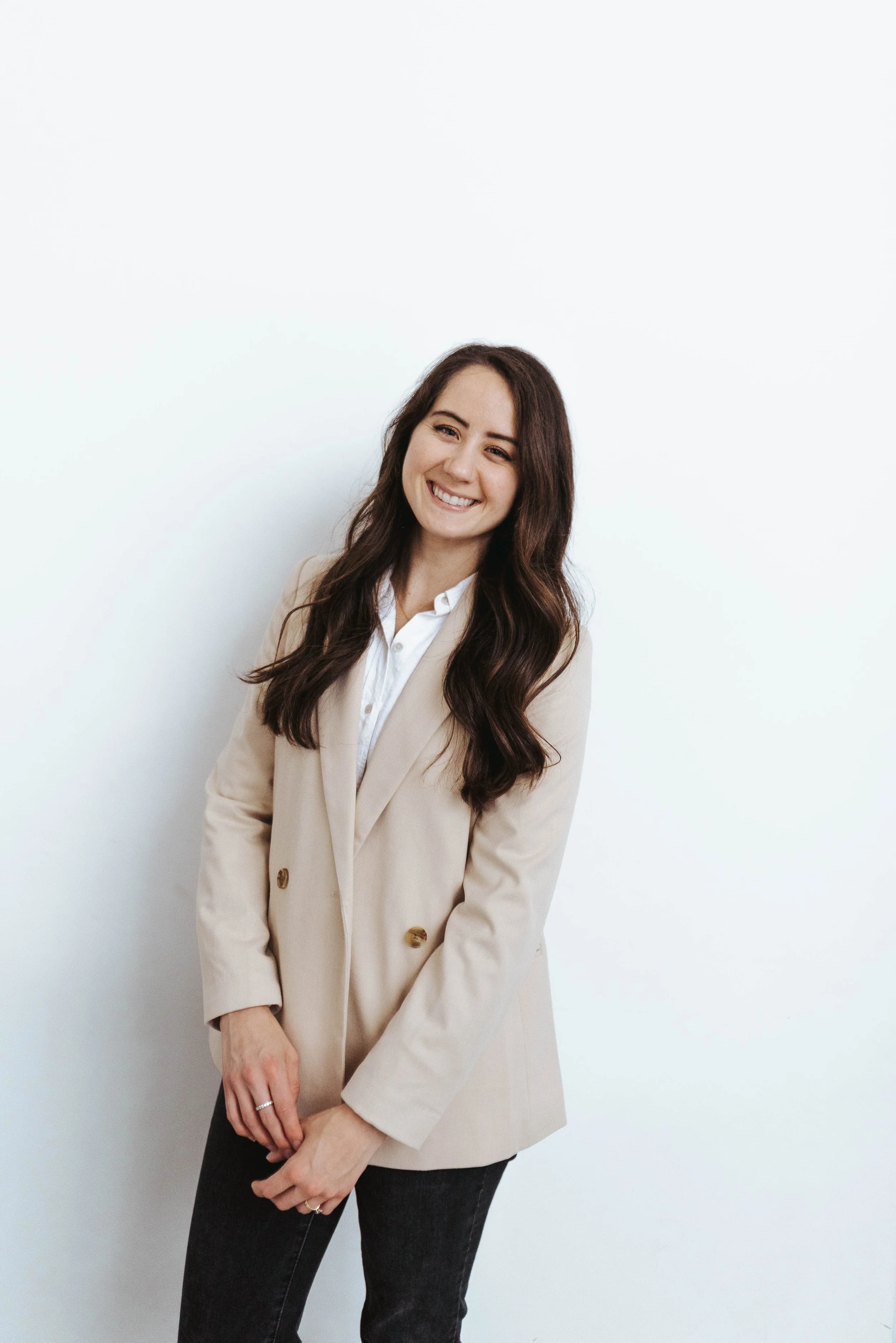 A woman with long brown hair styled in loose waves, smiling and wearing a beige blazer over a white shirt, standing against a plain white background.