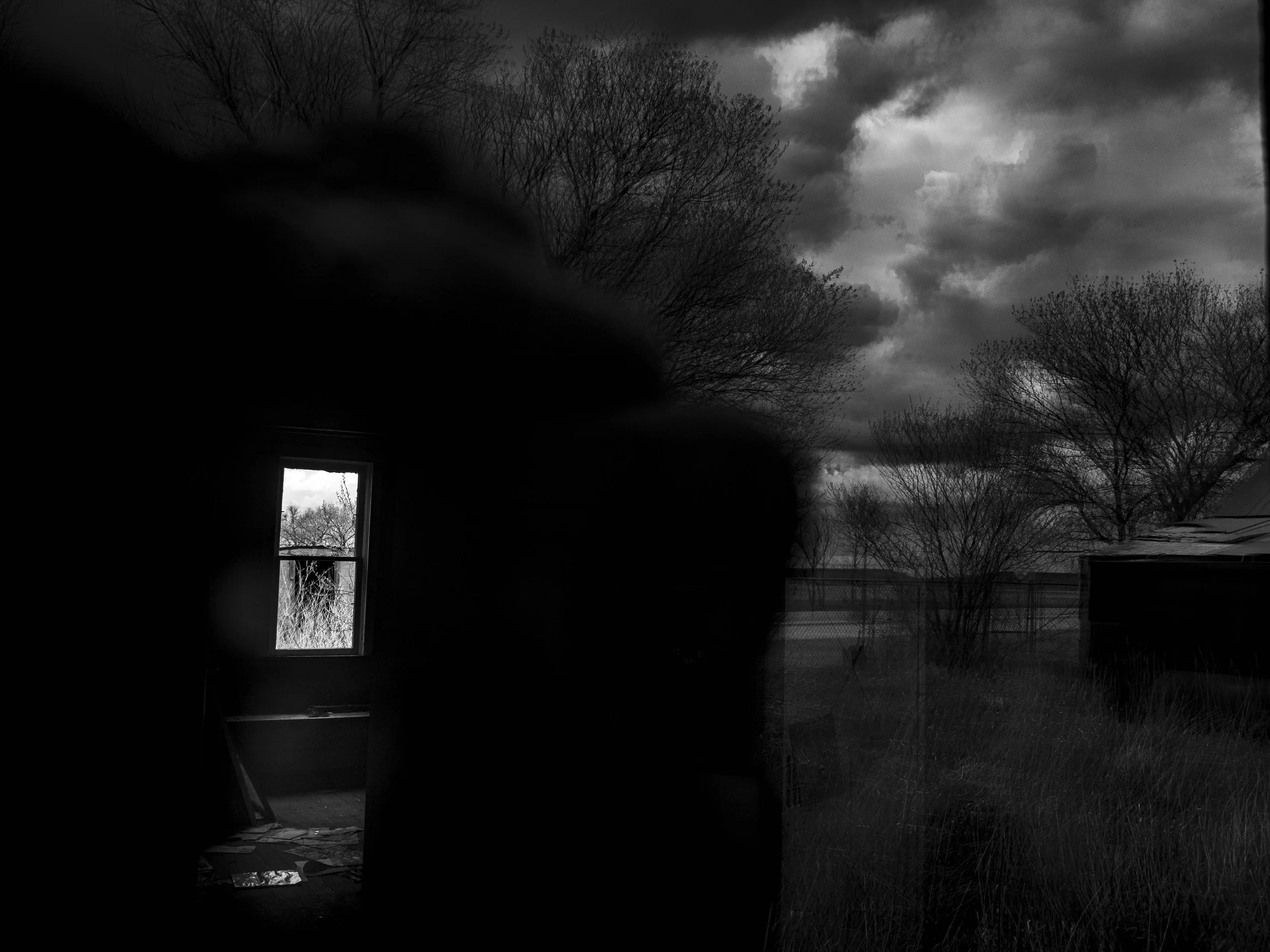 An abandoned room with a window showing an outside landscape of trees and a cloudy sky, with the interior in darkness.