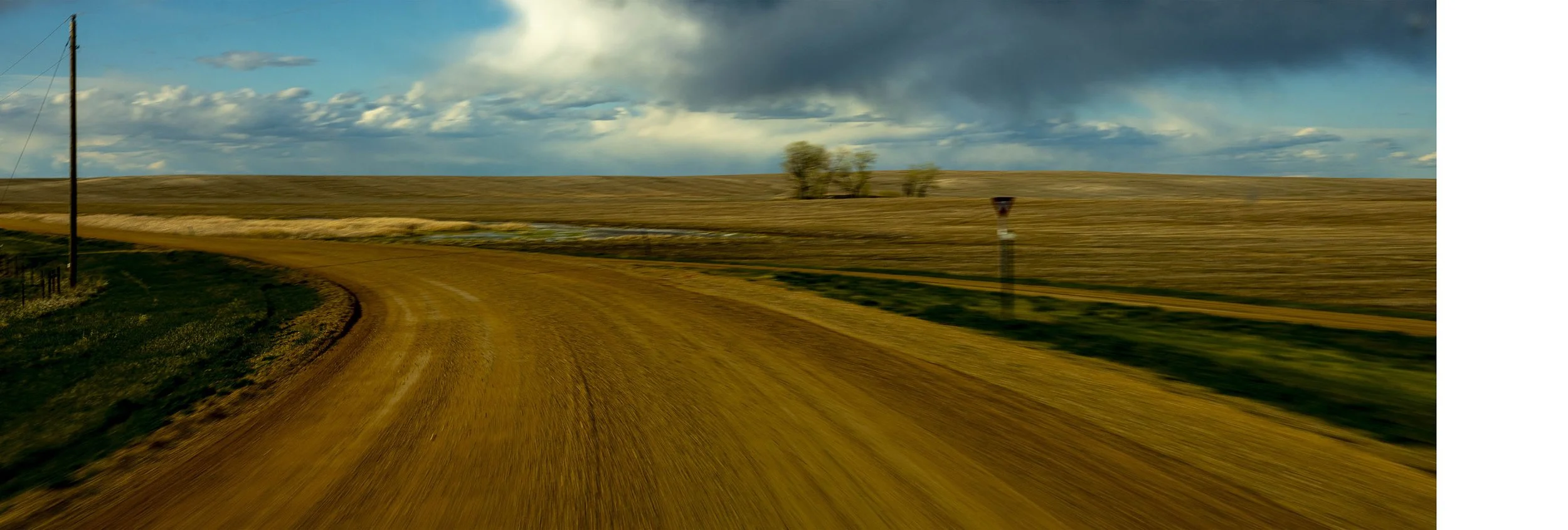 Rural dirt road curving through open farmland with a few trees and cloudy sky in the background.