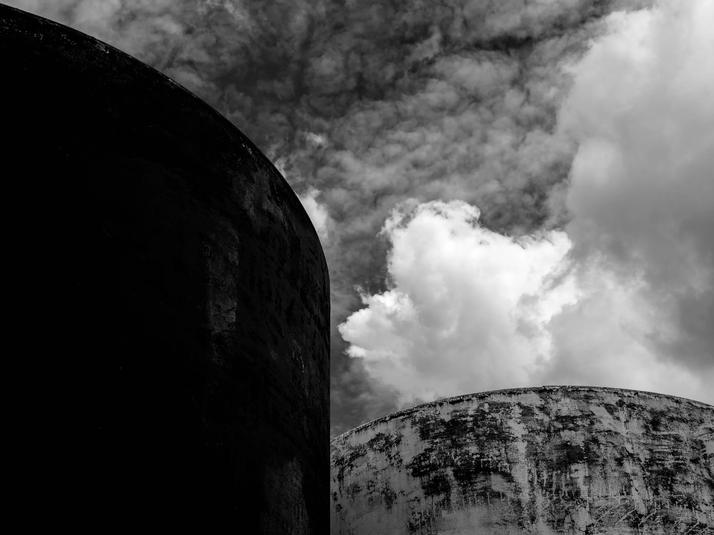 Black and white photograph of two large cylindrical tanks with weathered surfaces, set against a cloudy sky.
