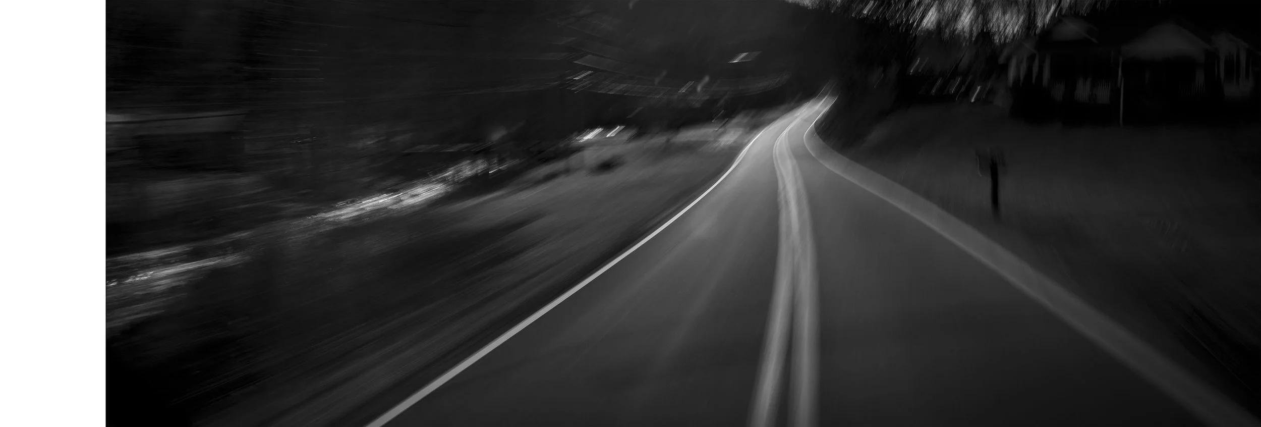Black and white photograph of a winding rural road at night, with trees on the sides and no visible vehicles.