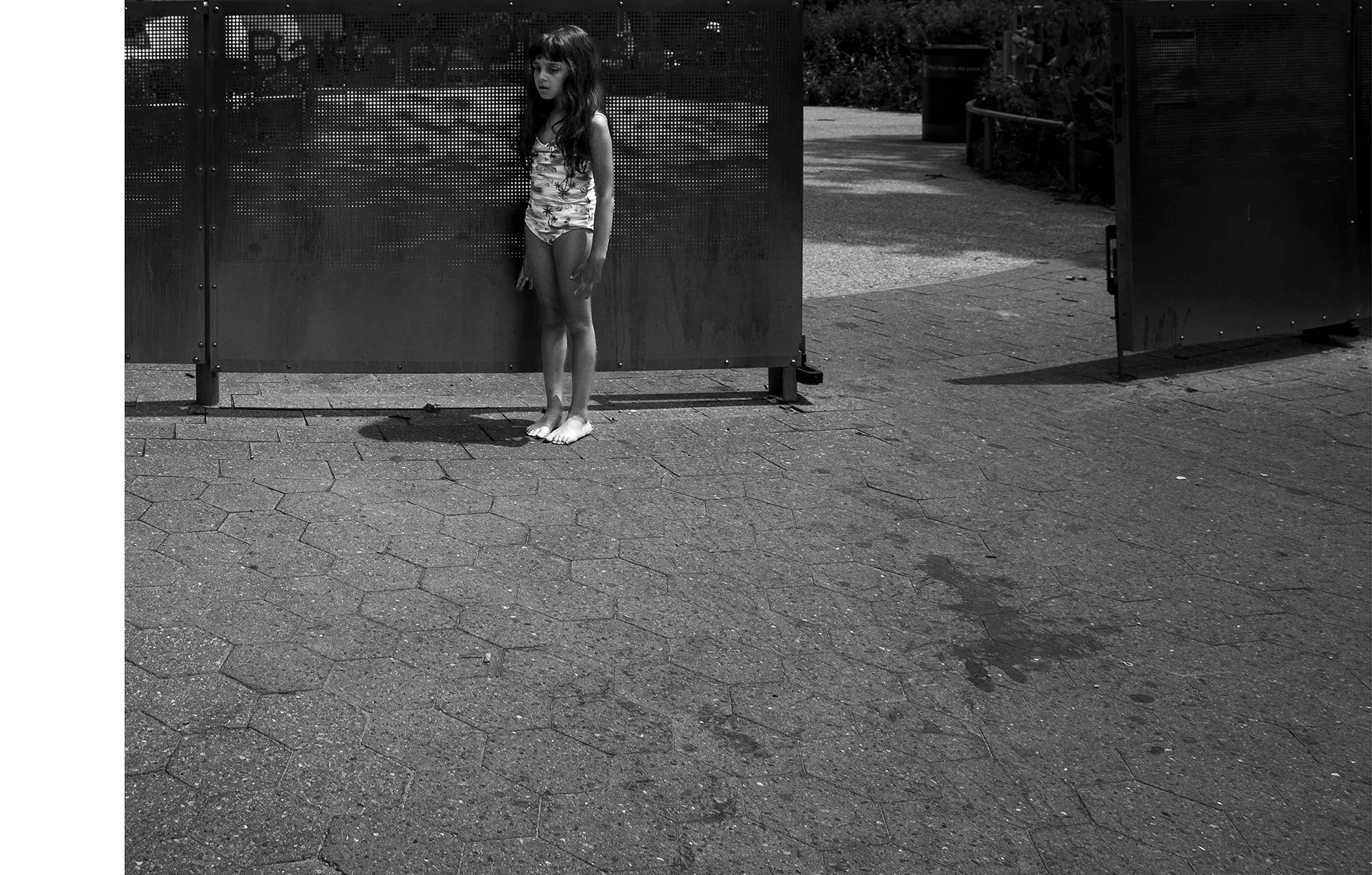 A young girl wearing a swimsuit standing barefoot on a paved sidewalk against a large metal fence or panel. The background includes some greenery and sidewalk area.