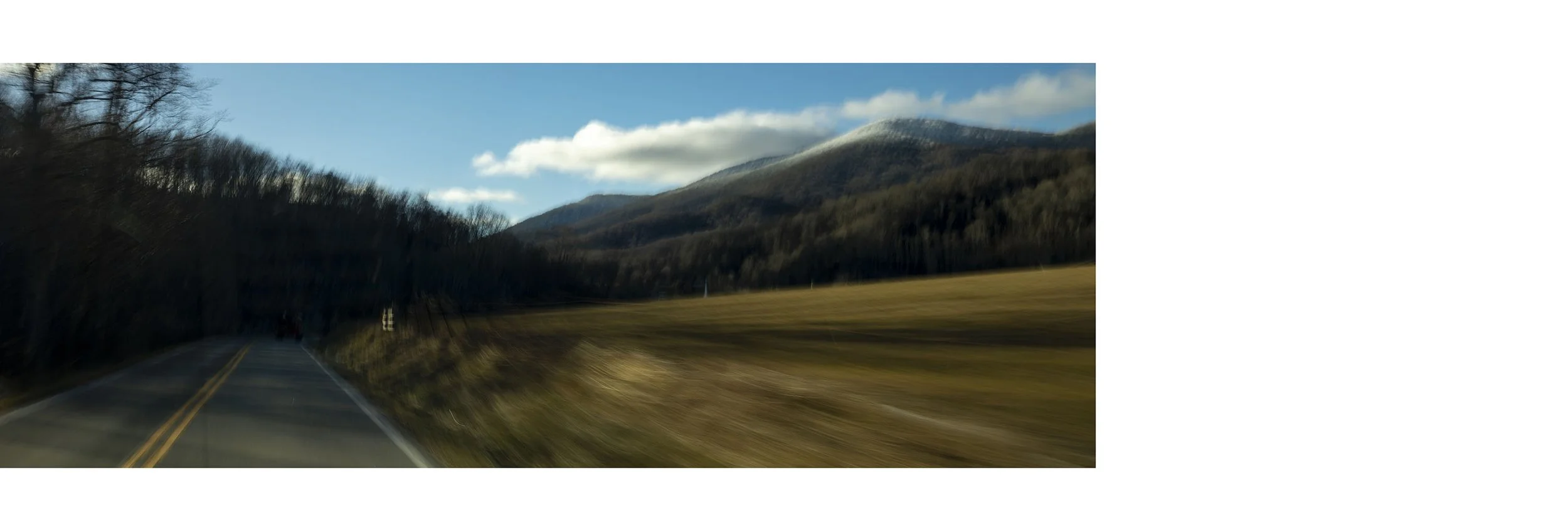 A scenic view of a two-lane road running through a rural area with mountains in the background and a partly cloudy sky.