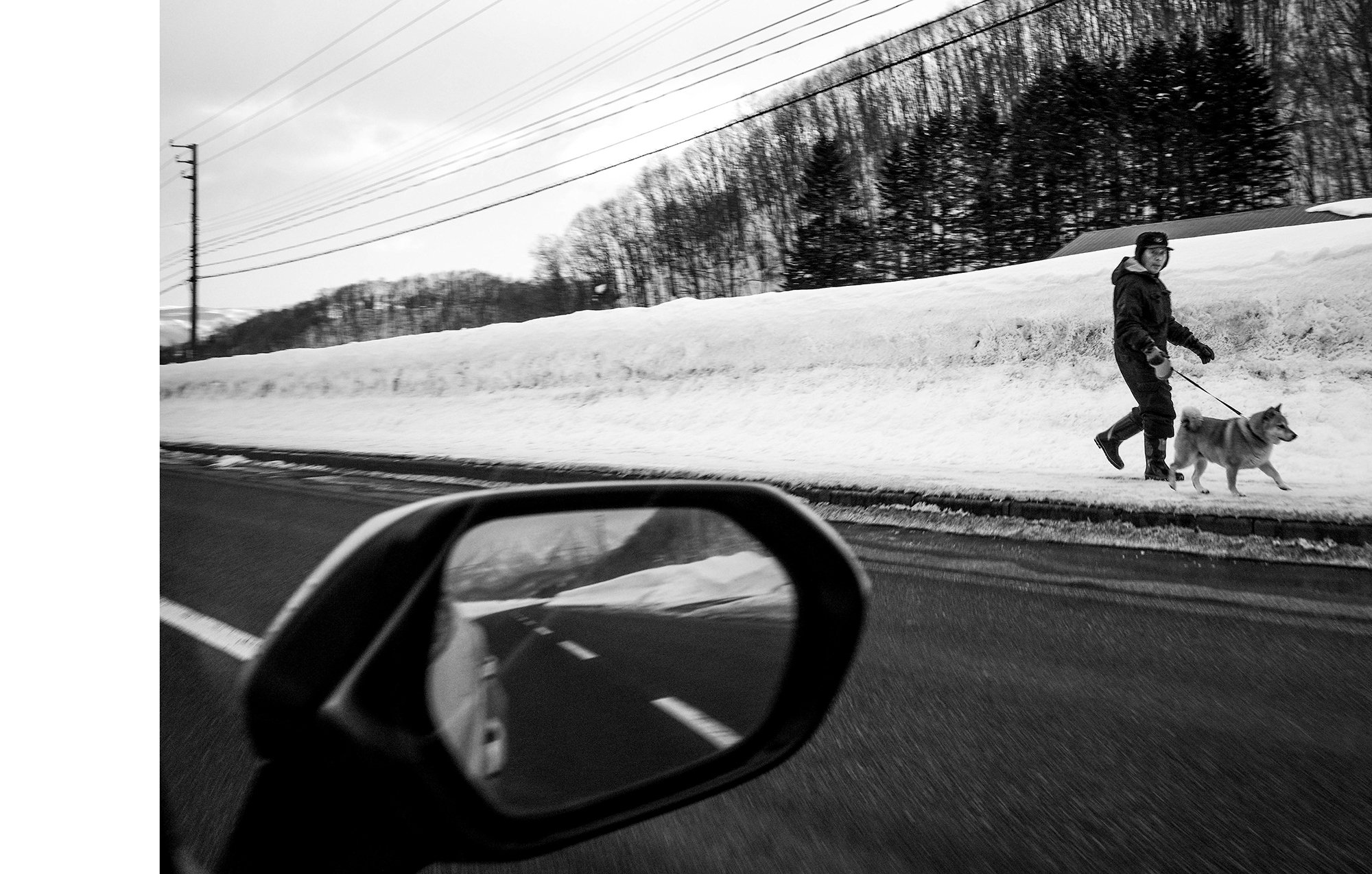 Black-and-white photo taken from inside a vehicle showing a person walking a dog on snow-covered sidewalk, with power lines, trees, and hills in the background.