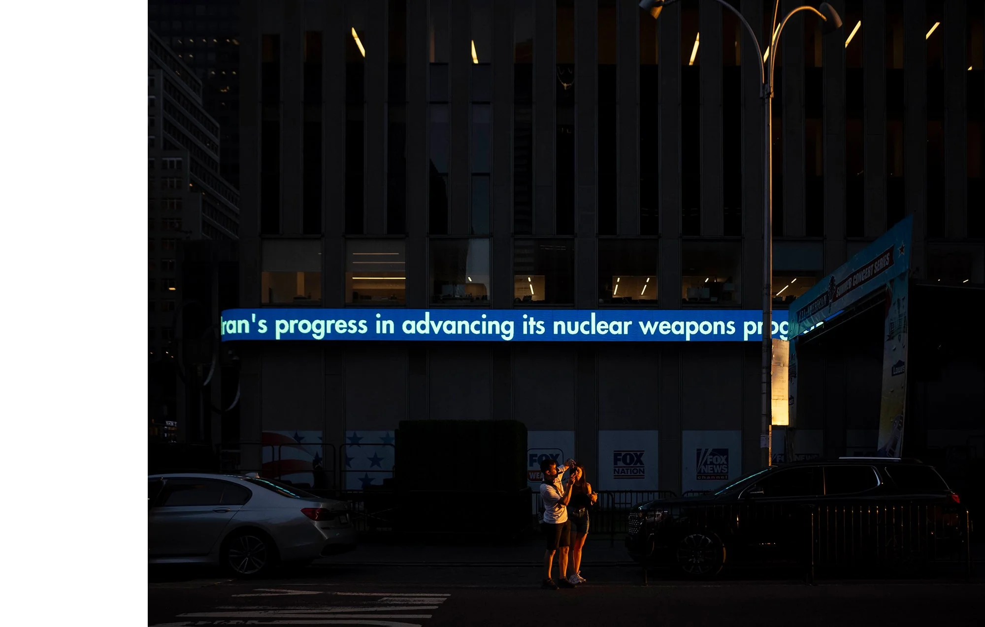 Two people standing on the sidewalk at night, taking a photo of each other, with cars parked behind them. Behind them is a building with a digital marquee displaying a partial news headline about nuclear weapons progress. The building has large glass