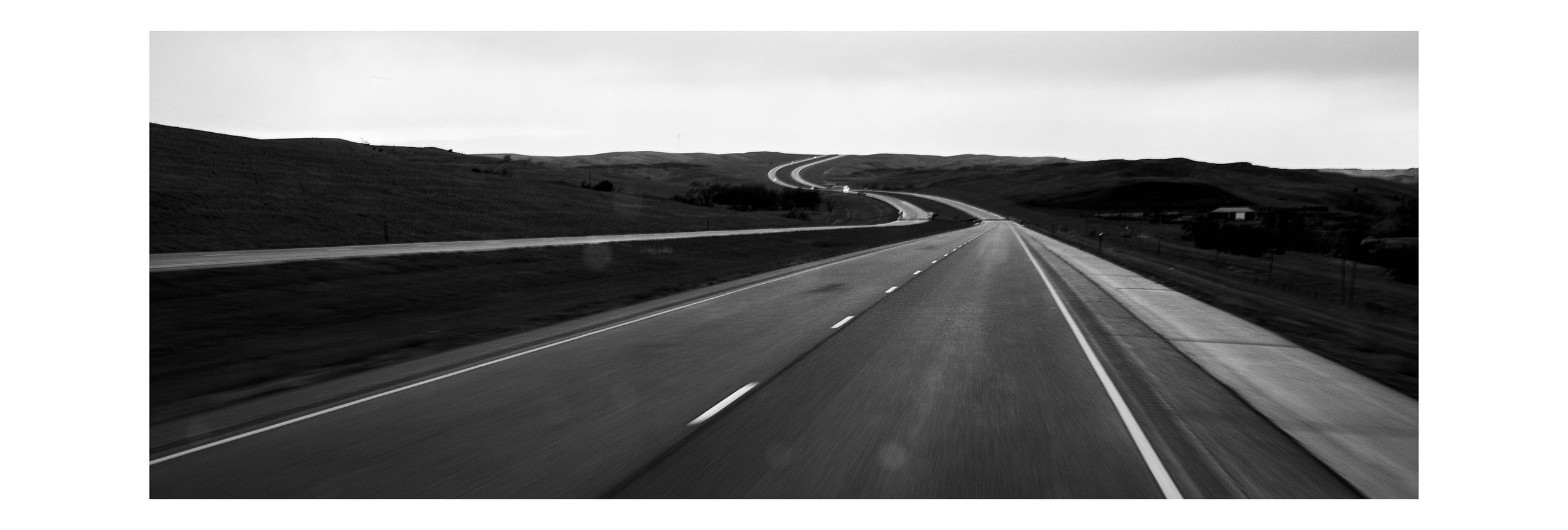Black and white photo of a winding highway stretching through rolling hills under an overcast sky.