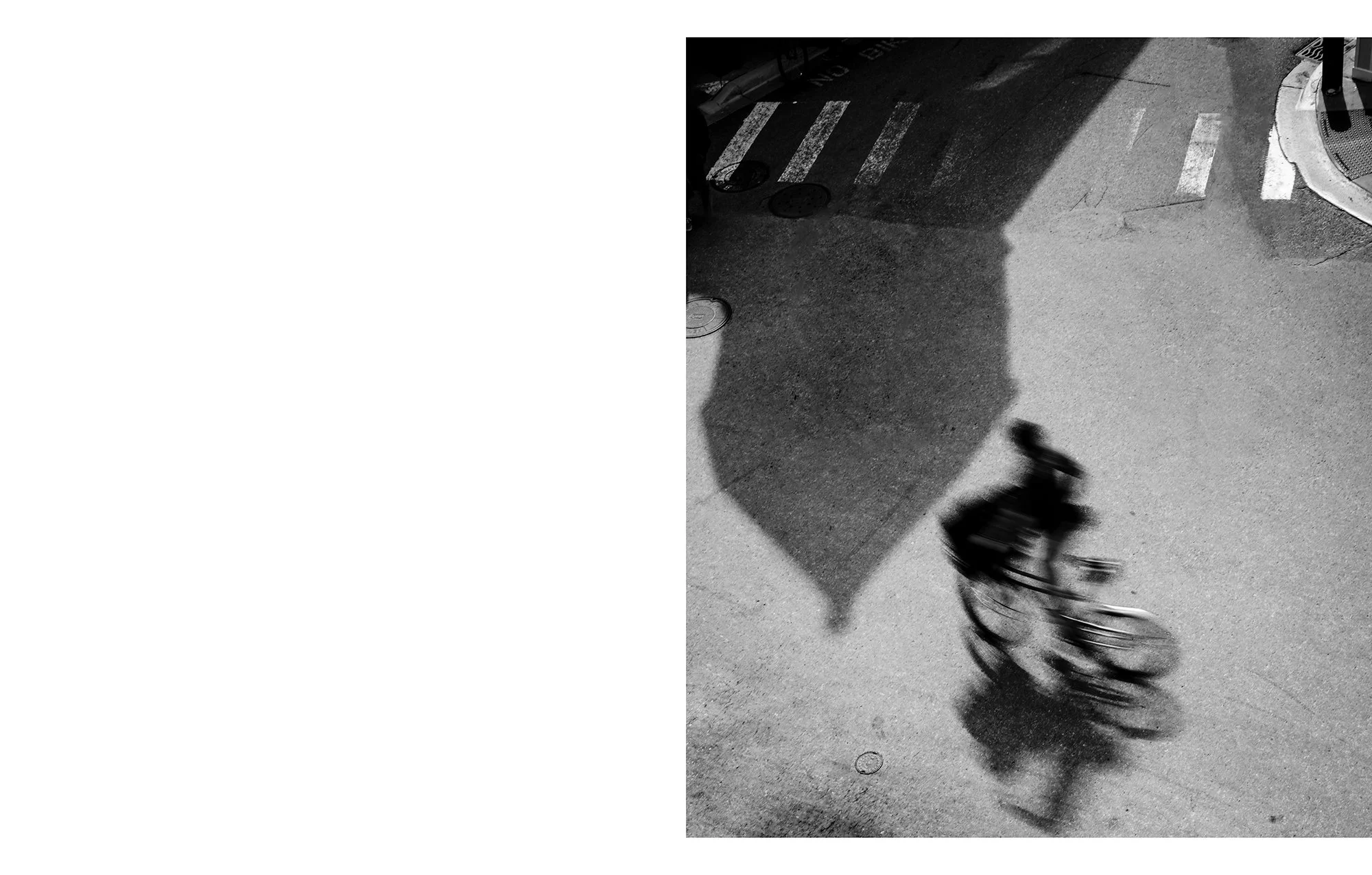 A shadow cast by an umbrella and a person riding a bicycle on a city sidewalk with markings and a curb.