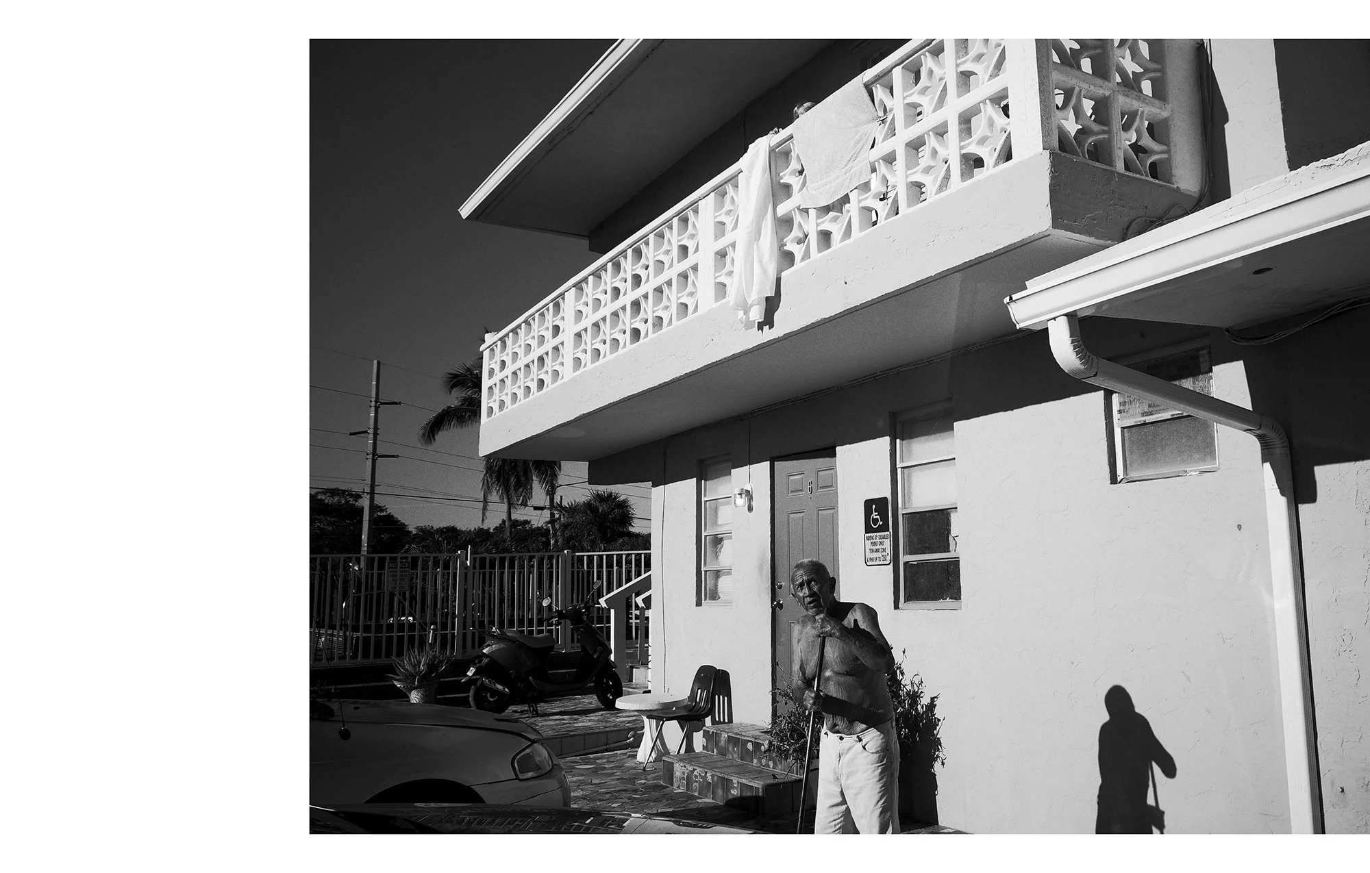 A black and white photo of a man with a cane standing outside a building. The building has a balcony with laundry hanging on the railing, a handicap sign, and a small car and scooter parked nearby.