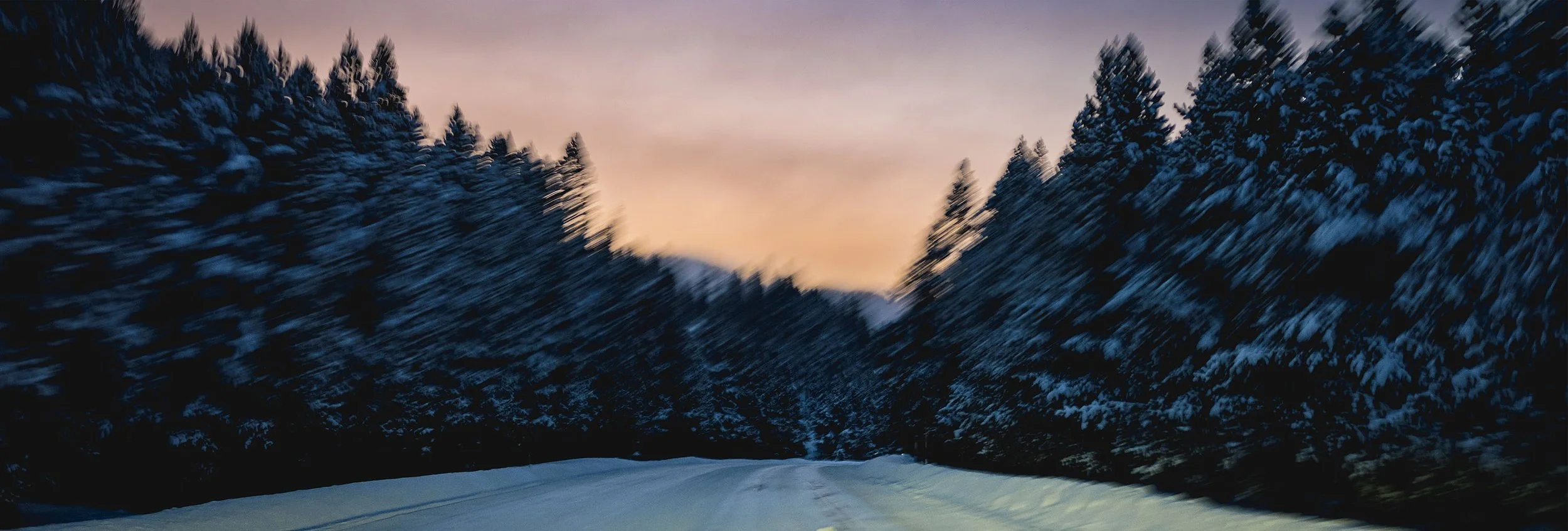 Snowy road surrounded by dark evergreen trees, with a colorful sunset sky in the background.