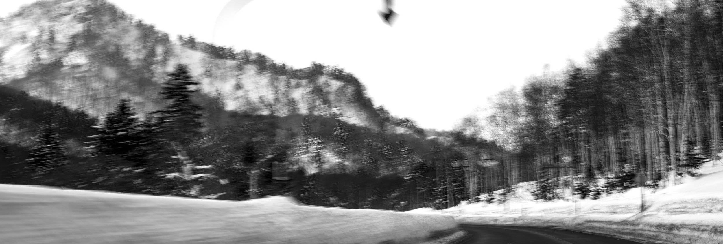 Black and white photograph of a winding mountain road surrounded by trees, with high rocky cliffs in the background.