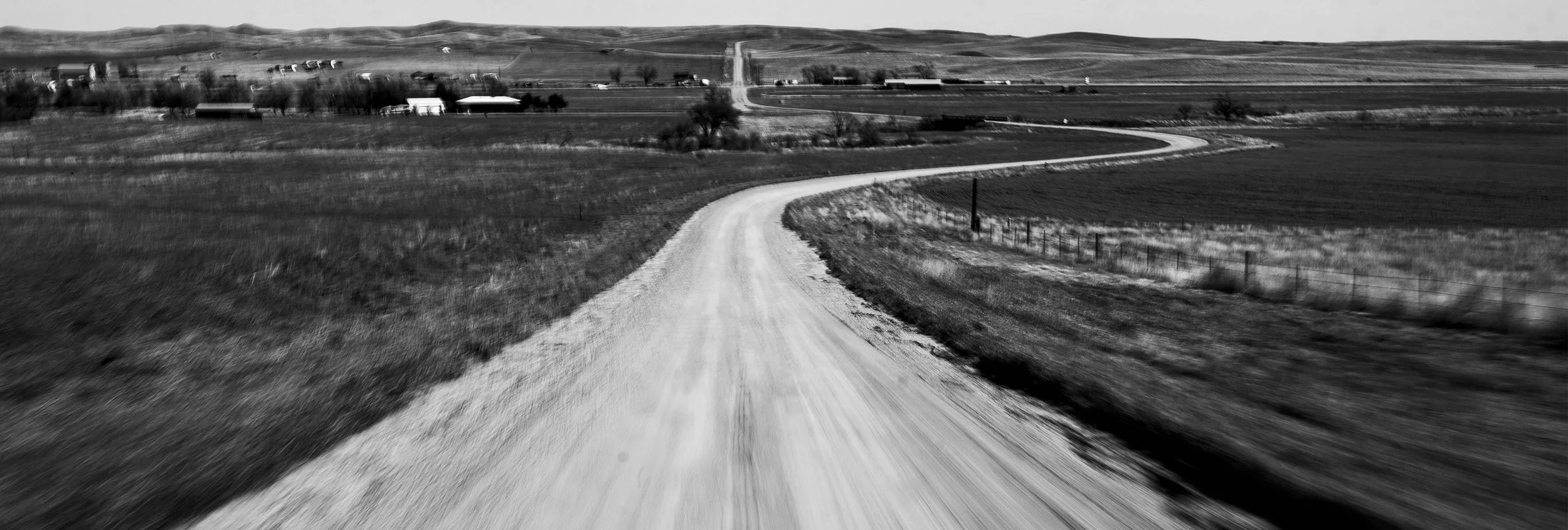 Black and white photo of a winding dirt road extending into rural farmland with houses and hills in the background.