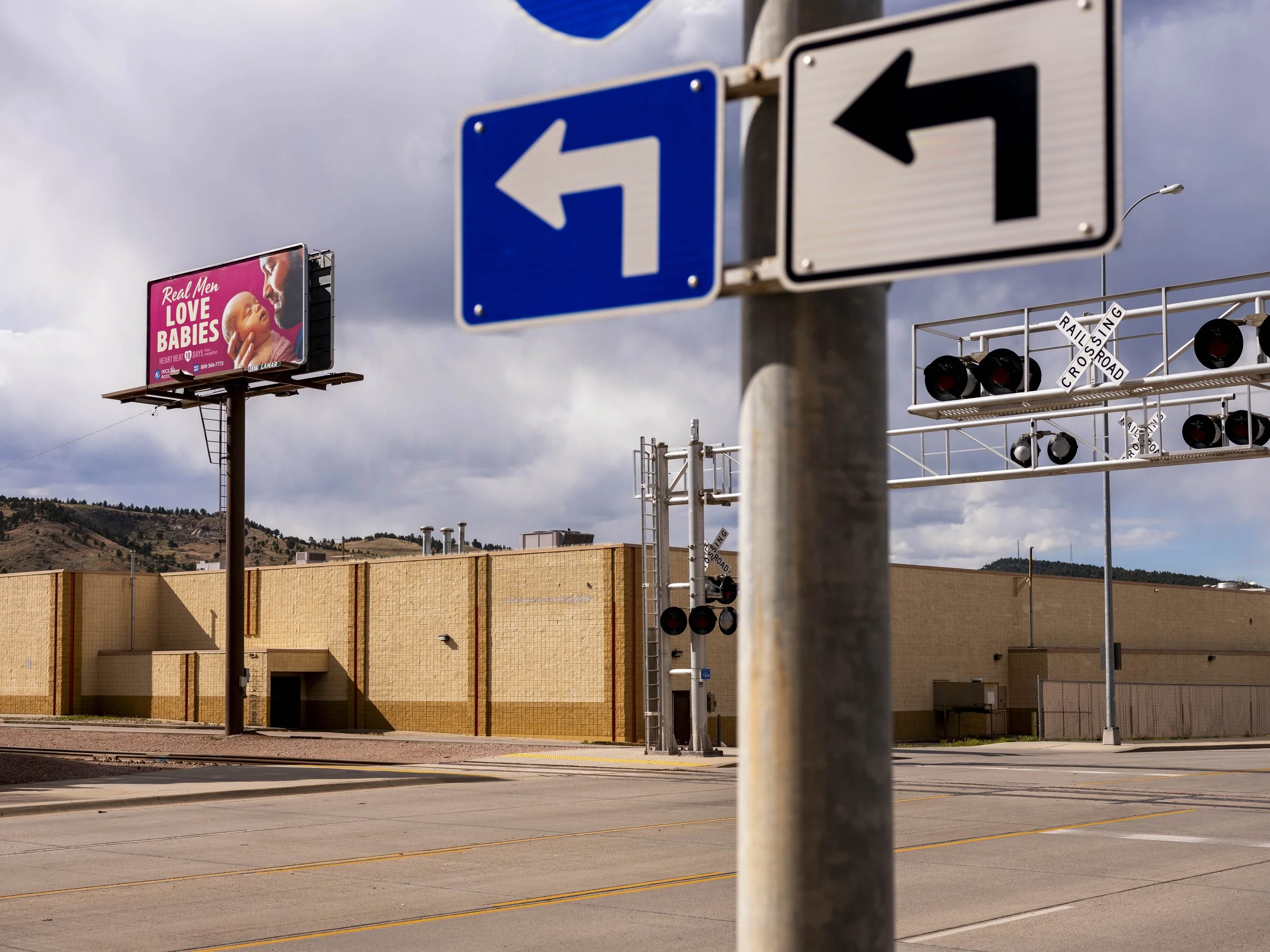 Street scene with traffic signs, railroad crossing signals, and a billboard showing a baby and a woman with the text "Real Men Love Babies."