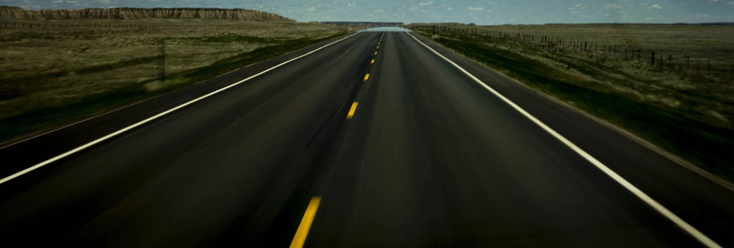 A long, straight road stretching through a rural landscape with grassy fields and low hills, under a partly cloudy sky.