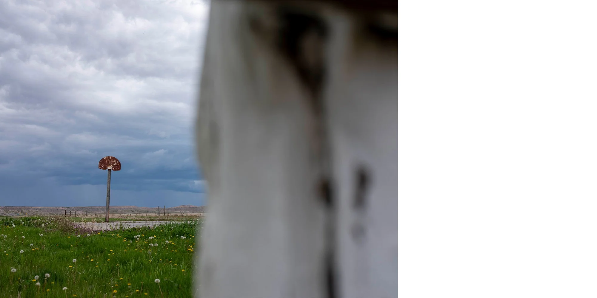 A rusty, half-sunken basketball hoop standing on a grassy field under a cloudy sky, with a blurred white wall or object partially blocking the right side of the view.