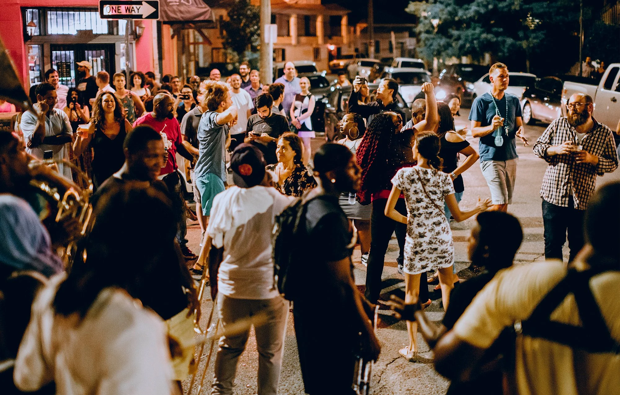 A diverse group of people gathered on a city street at night, socializing, dancing, and enjoying the evening. There are cars parked along the street, and a 'One Way' sign is visible. The scene is lively with various individuals engaging and taking ph